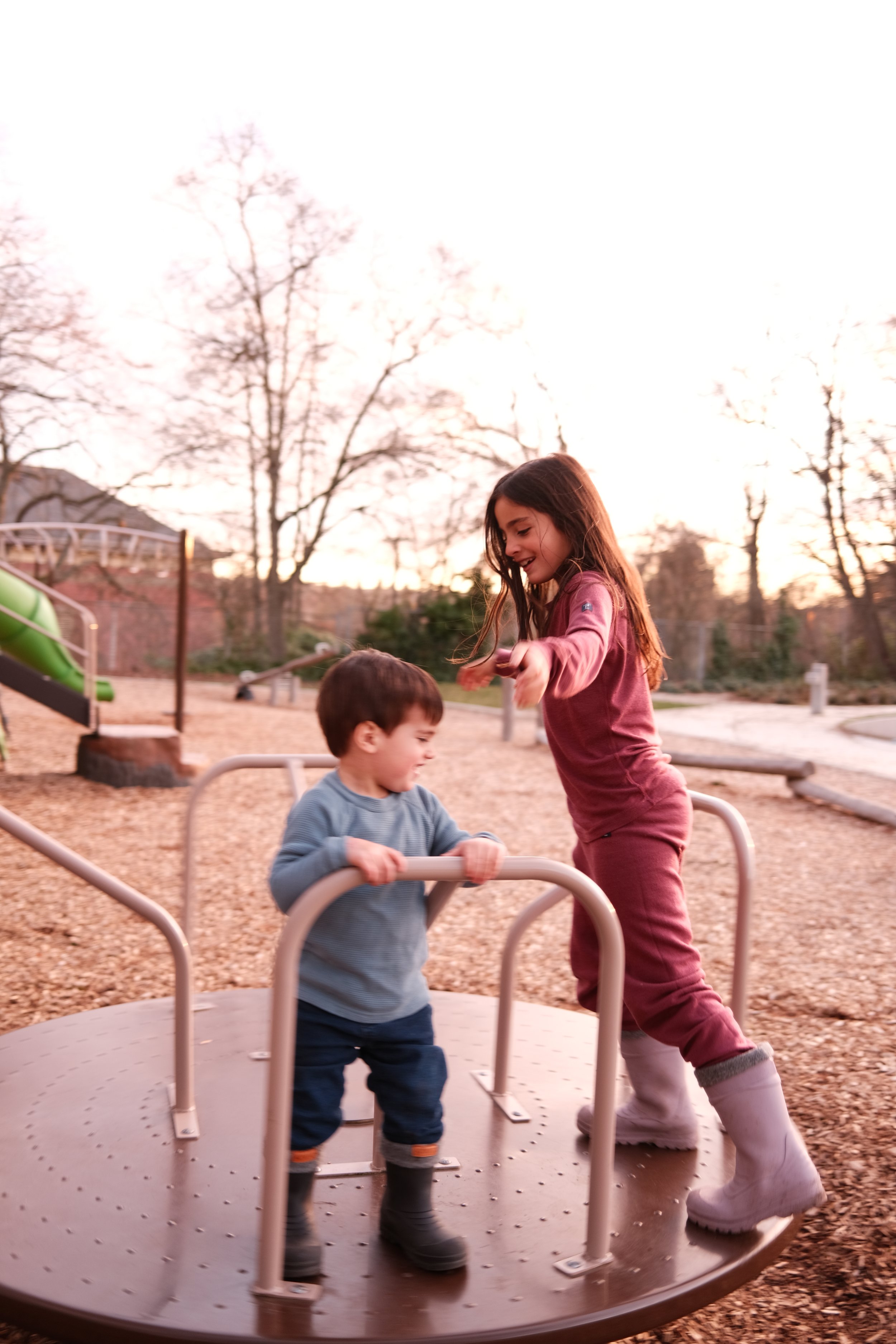 Crianças brincando em um parque ao pôr do sol, uma criança mais velha ajudando uma mais nova em um brinquedo de rodar.