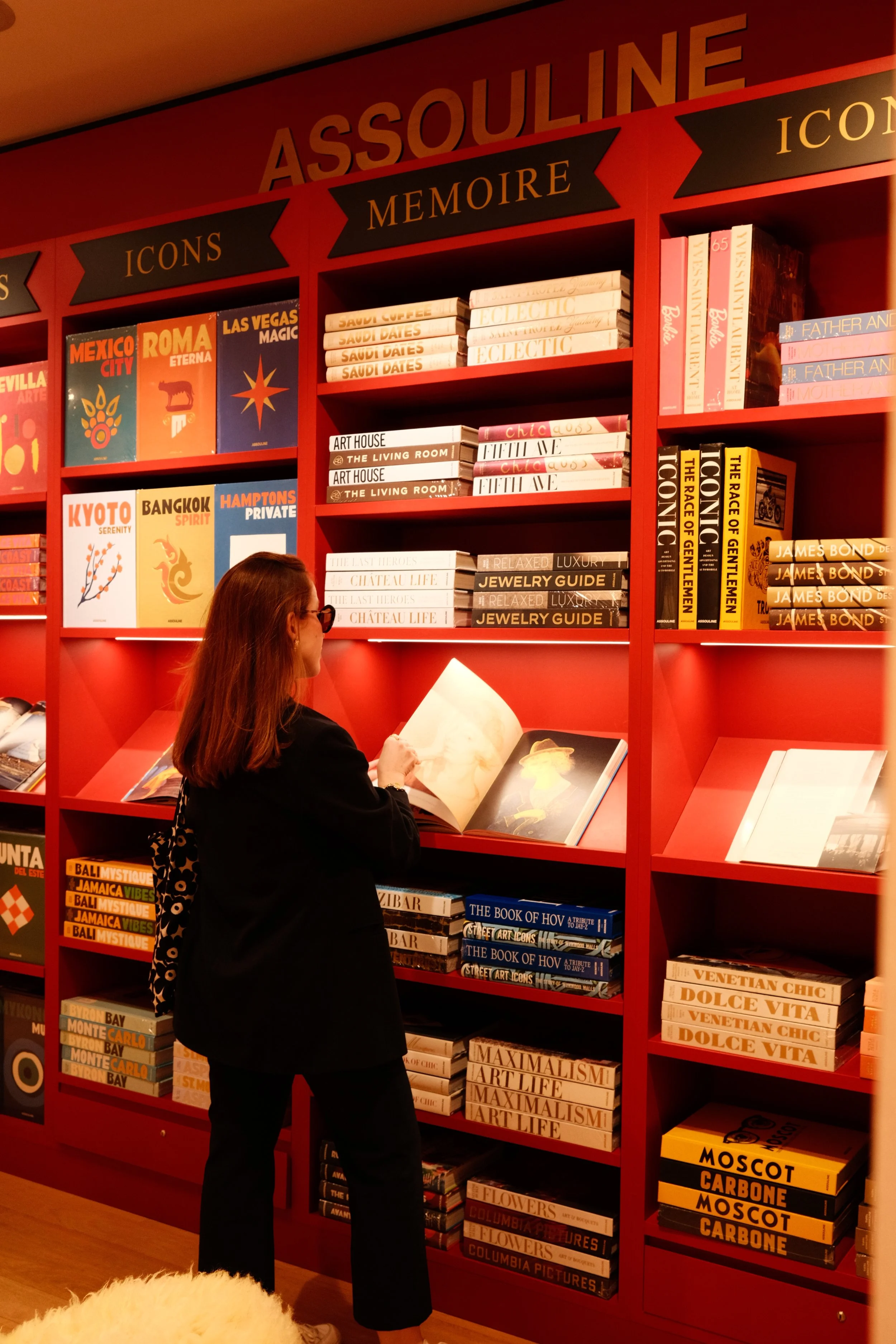 Pessoa lendo livro em uma estante de cores vermelhas na loja de livros Assouline, com várias prateleiras de livros de capa dura e selo dourado.