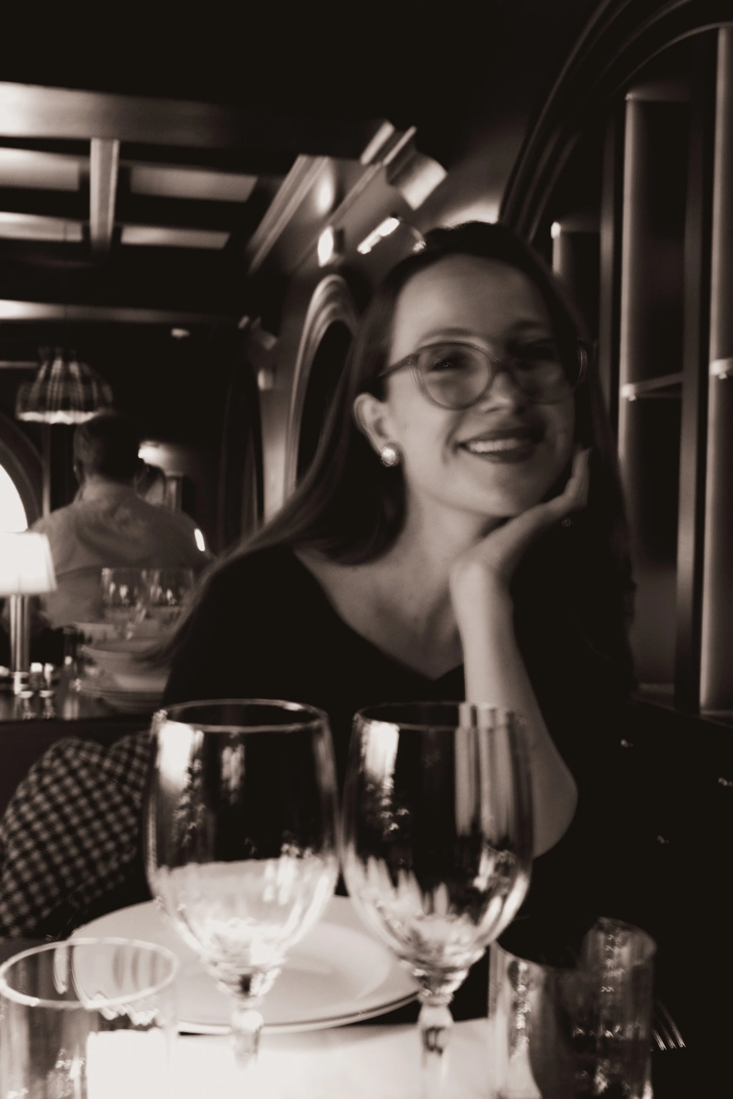 Mulher sorrindo, de óculos, sentado a uma mesa de restaurante com taças de vinho na frente, ambiente de jantar em preto e branco.