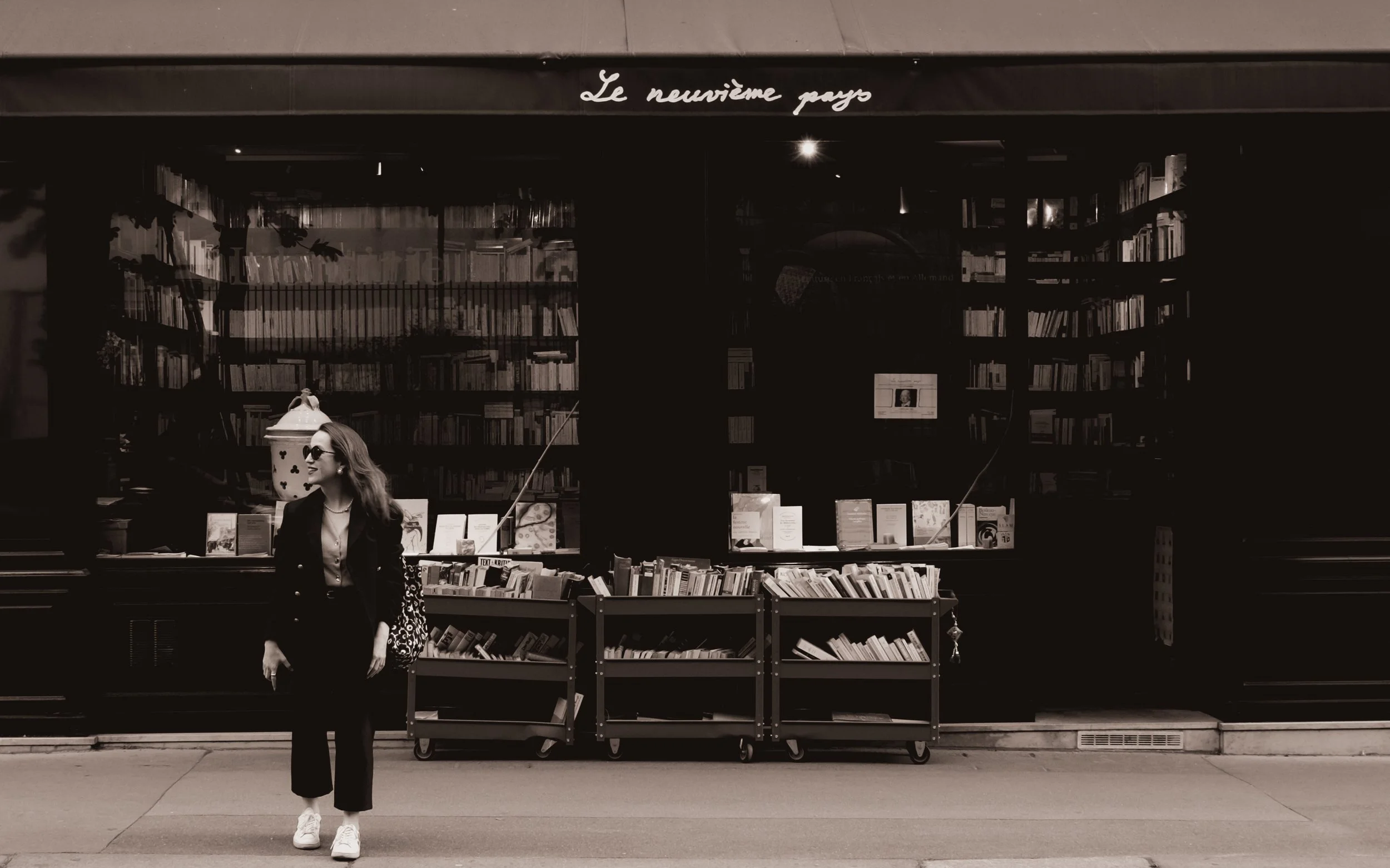 Pessoa sorrindo na calçada na frente de uma loja de livros com fachada de vidro cheia de livros e cartazes, escrita 'Le neuvième pays' na parte superior.