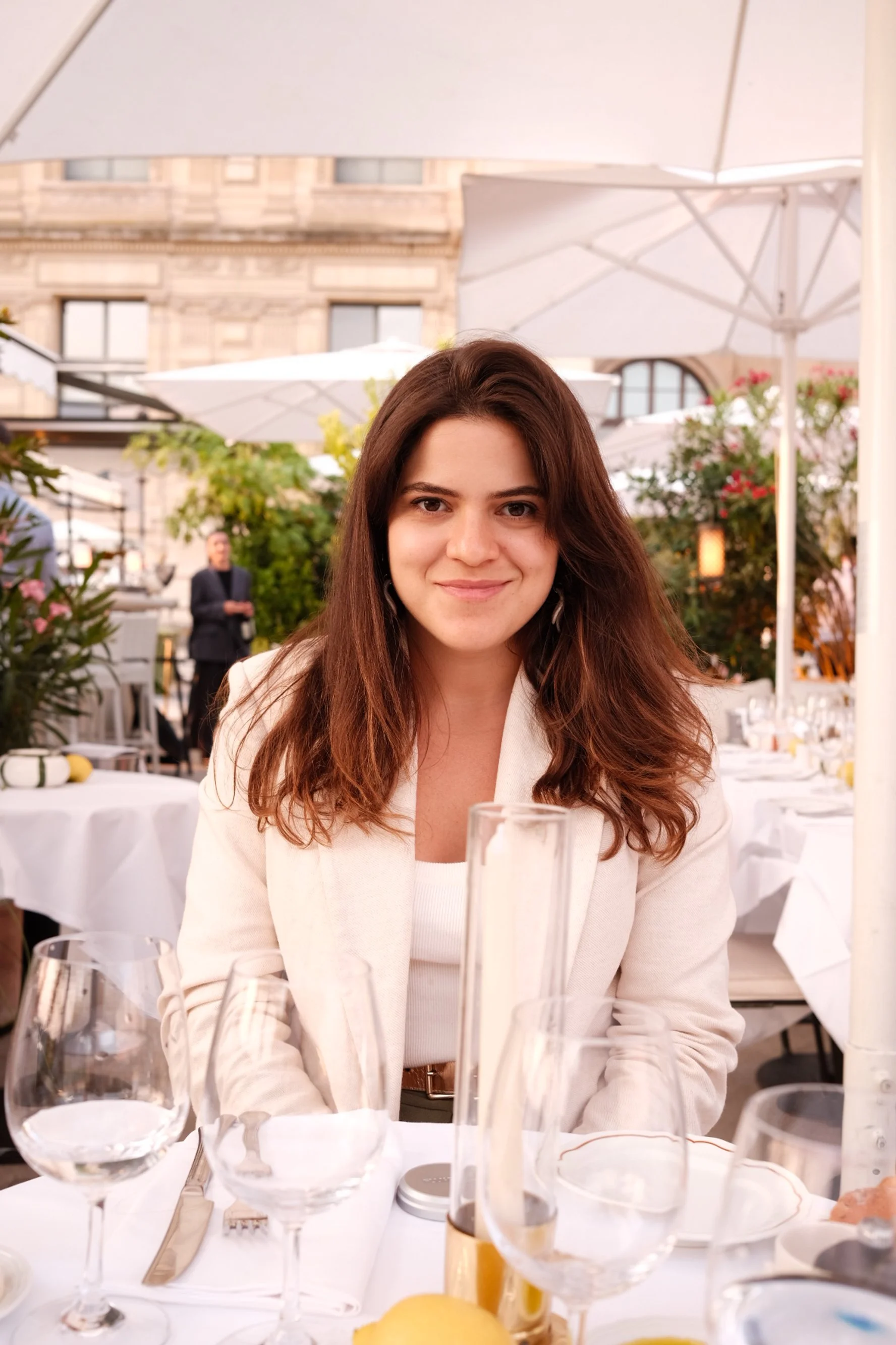 Mulher com cabelo castanho encaracolado sentada à mesa em restaurante ao ar livre, com taças de vinho e alimentos na mesa, sob guarda-sóis brancos, com edifício antigo ao fundo.