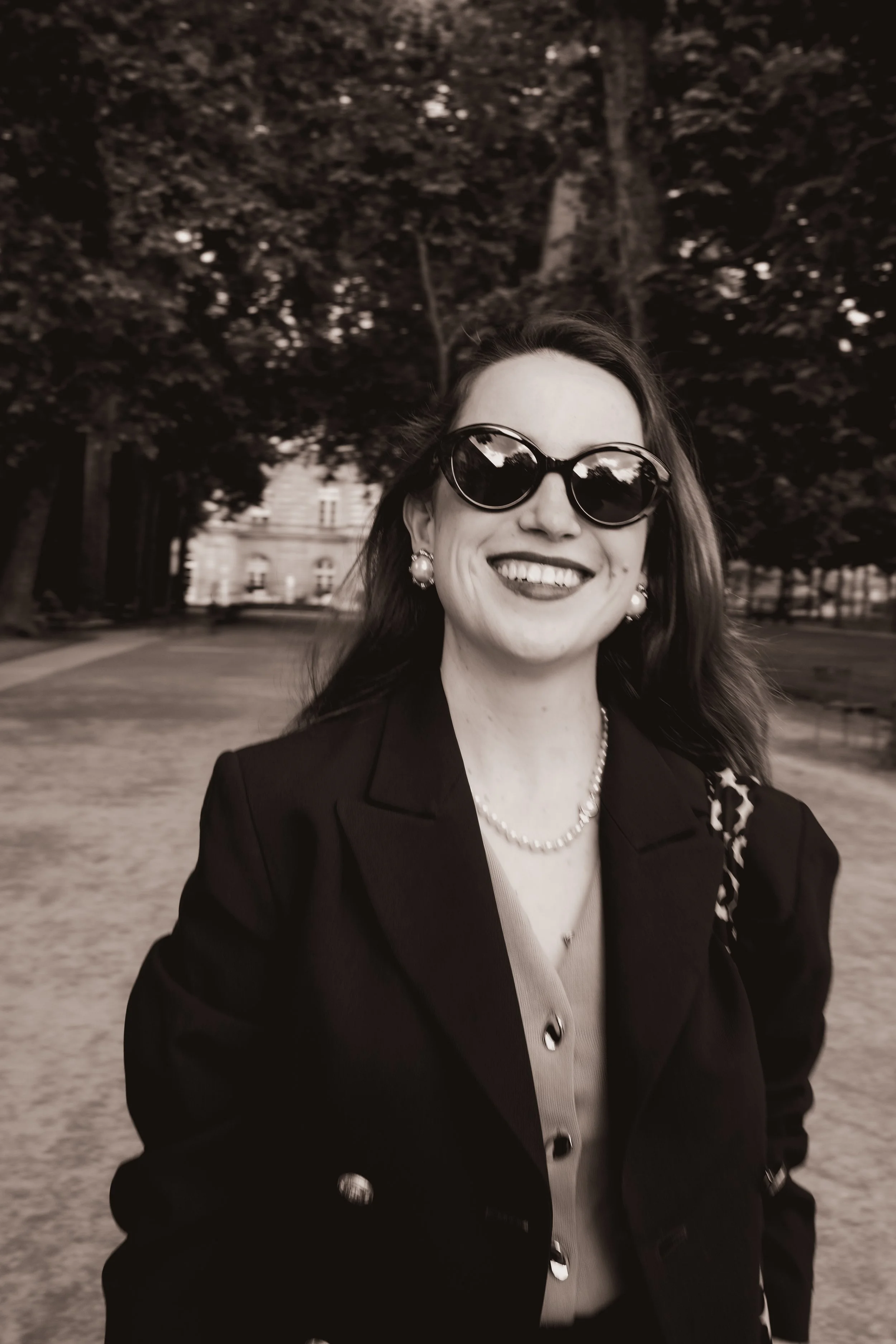 Mulher sorridente usando óculos escuros, brincos de pérola, colar de pérola, blazer escuro e camisa clara, ao ar livre sob árvores