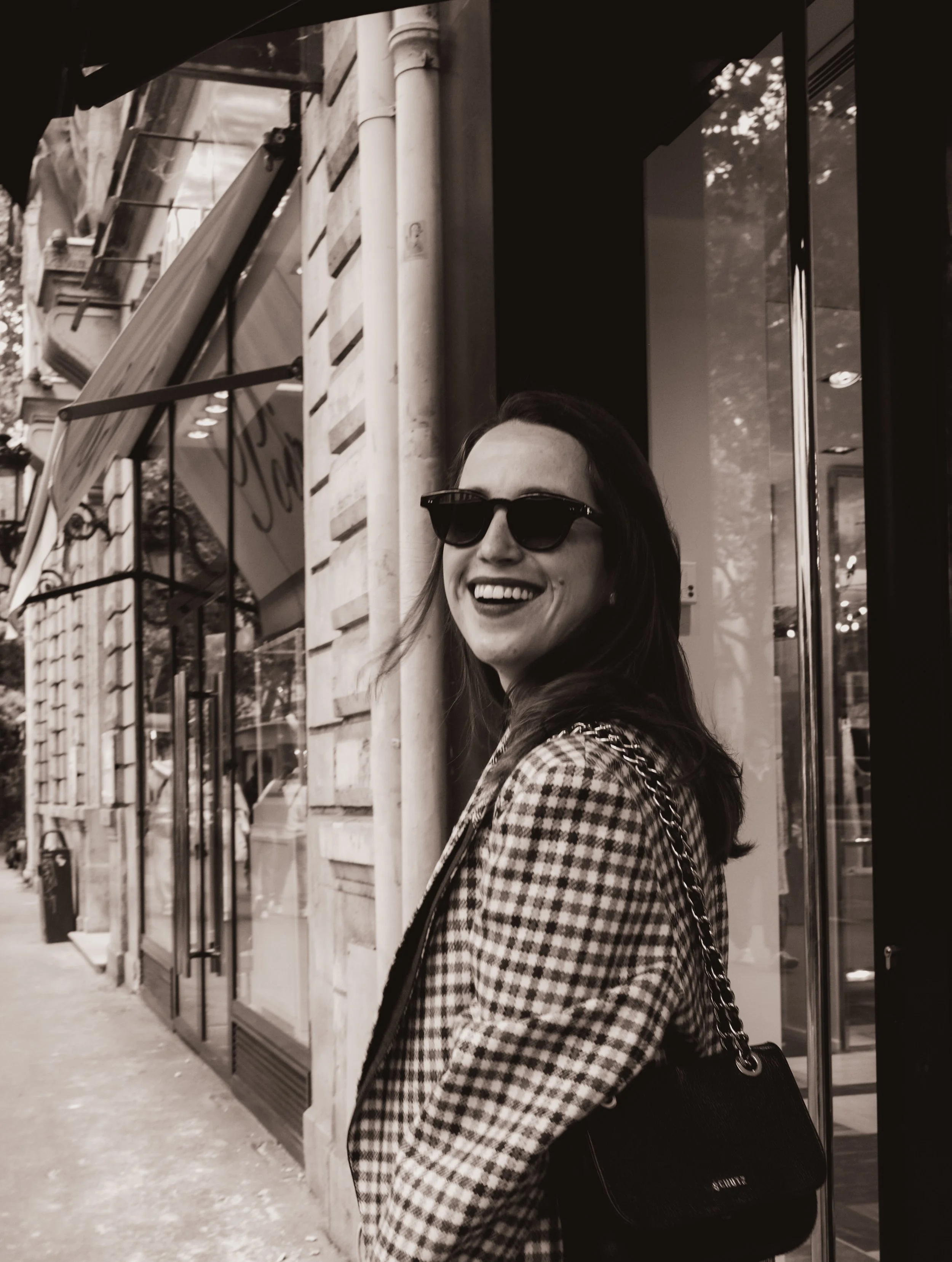 Mulher sorrindo usando óculos escuros e blusa de xadrez, na rua, ao lado de uma loja.