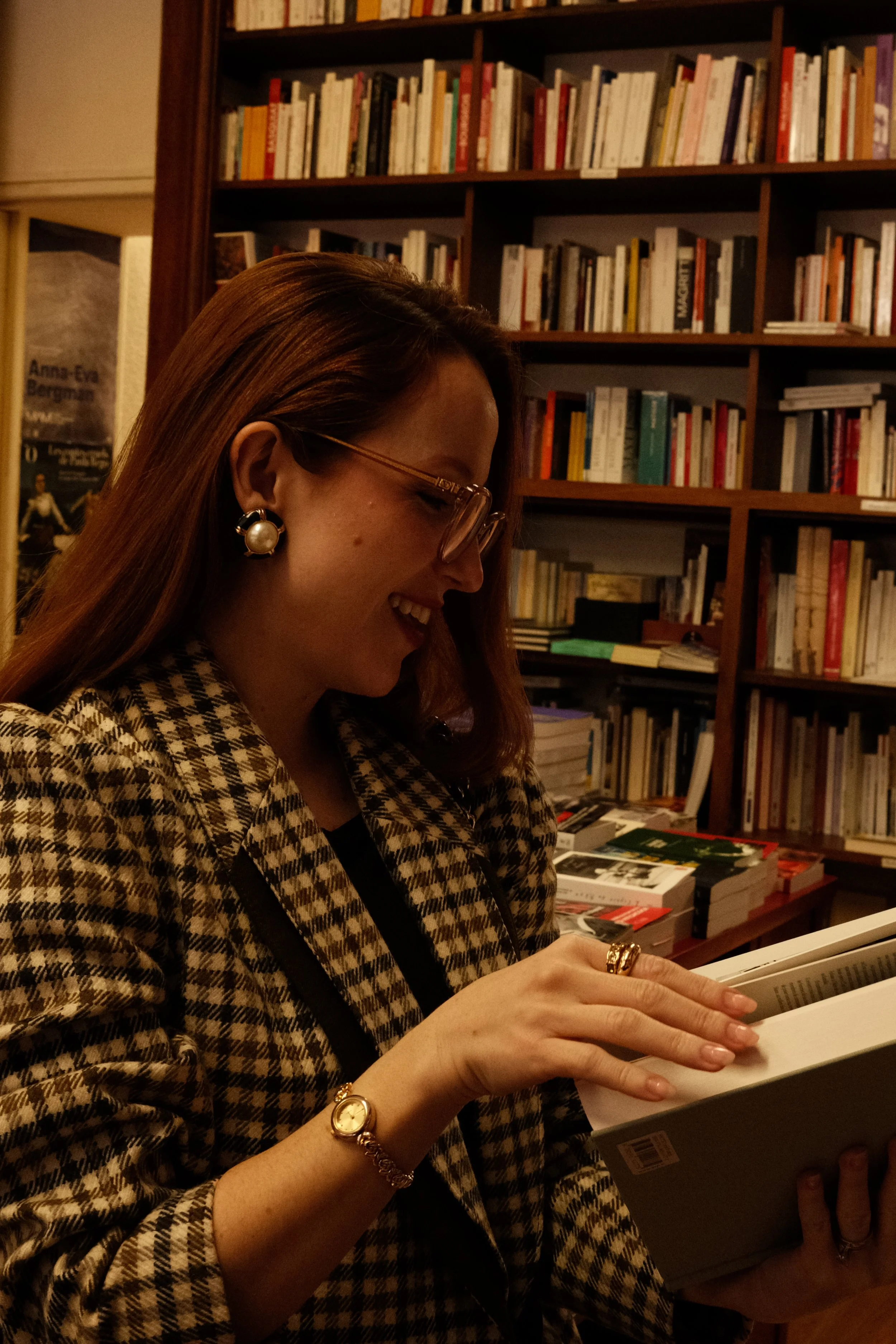 Mulher sorridente lendo um livro em uma biblioteca com estantes de livros ao fundo.