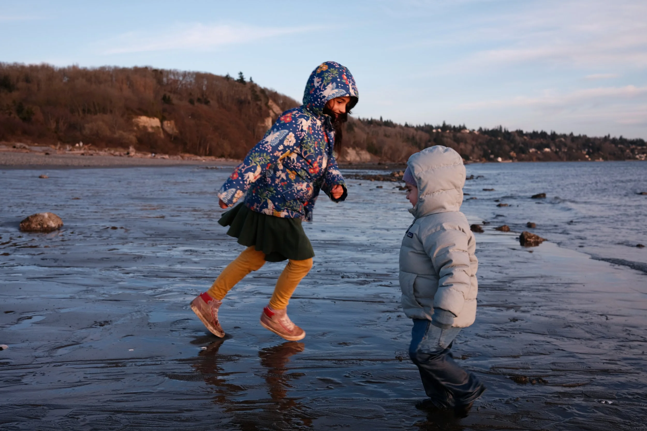 Duas crianças brincando na praia, uma pulando e a outra observando, com mar e colinas ao fundo, em clima de fim de tarde.