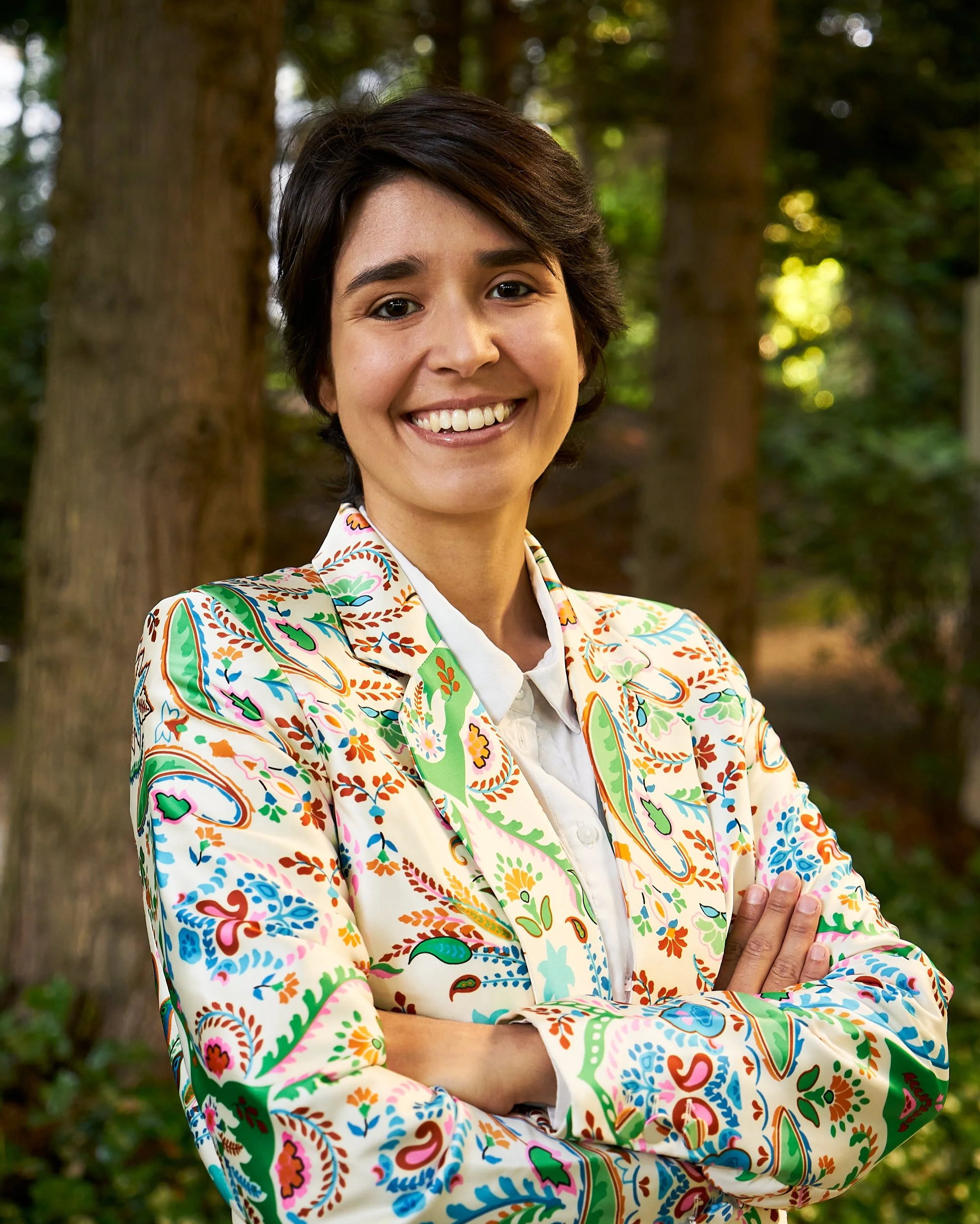 A person with short dark hair and a bright smile wearing a colorful, patterned blazer in front of trees with sunlight filtering through the leaves.