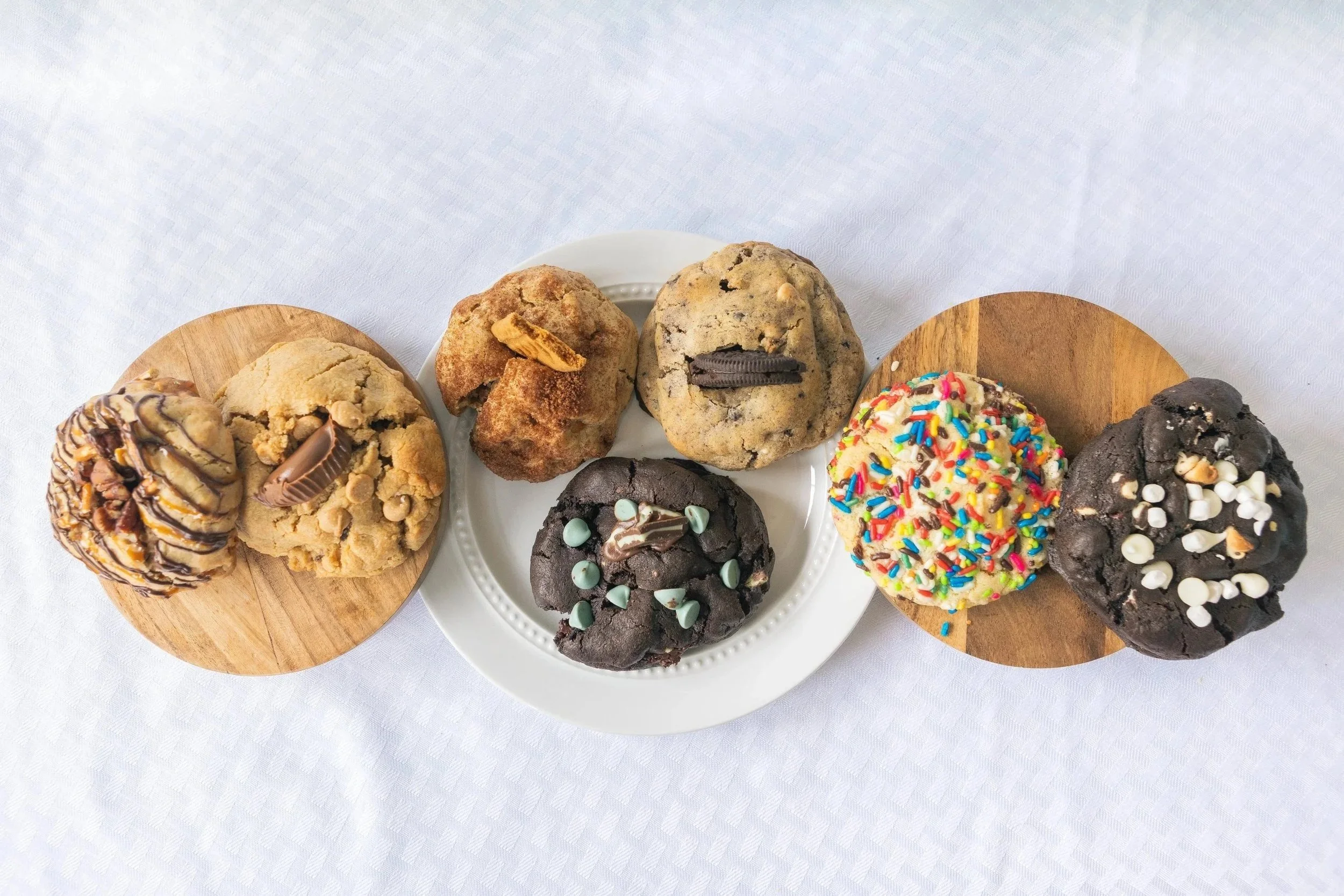 Assorted cookies on small wooden and ceramic plates, including chocolate chip, sprinkled, and decorated cookies, on a white tablecloth.