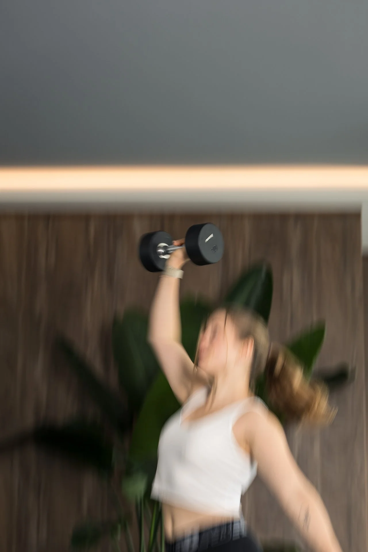 A woman in a white sleeveless top lifting a black dumbbell with her right hand in a fitness or workout setting.