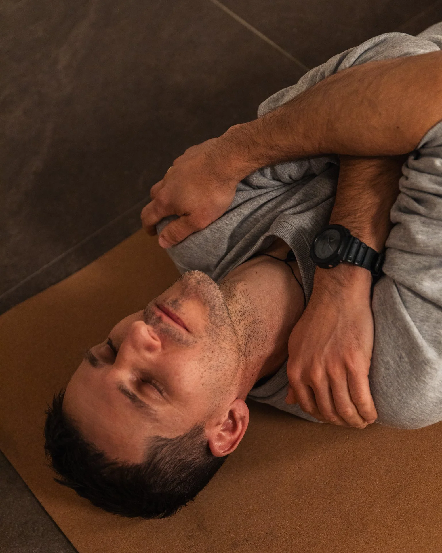A man lying on his side on a yoga mat, eyes closed, with arms crossed over his chest, wearing a gray shirt and a black watch.