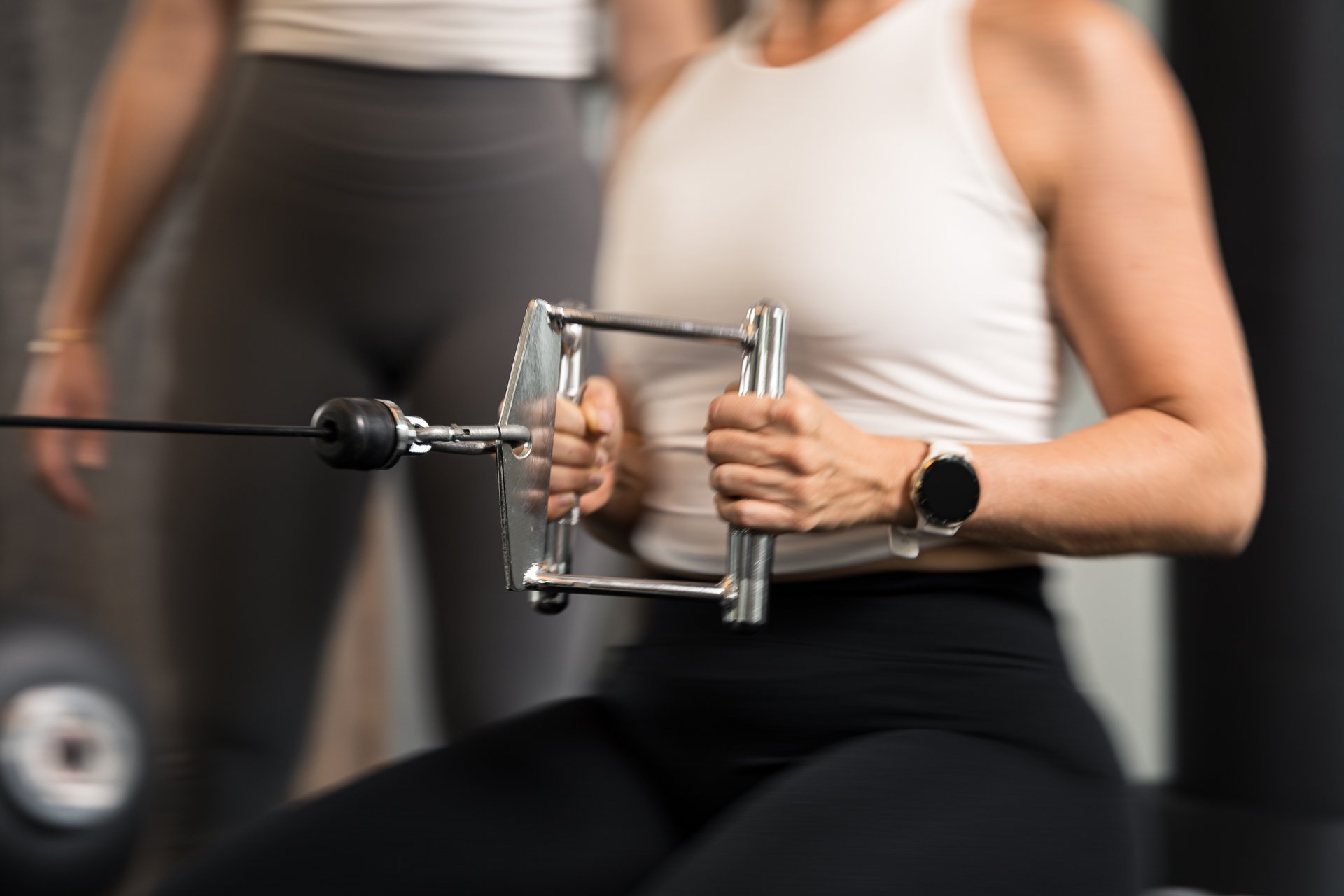 Woman using a rowing machine at gym, wearing a white tank top and black workout pants.