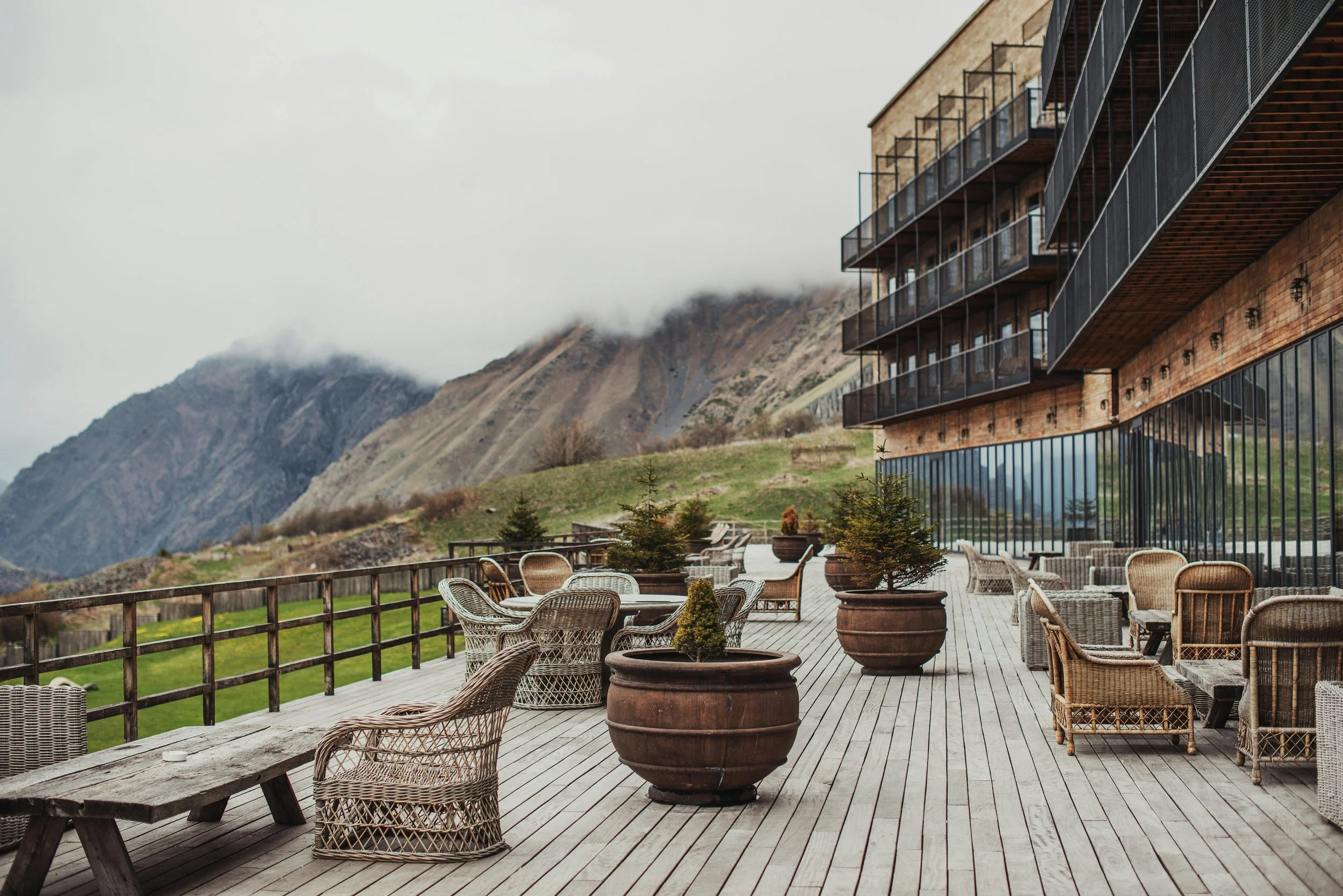 Empty outdoor terrace with wicker and wooden furniture, potted trees, and mountain scenery in the background.