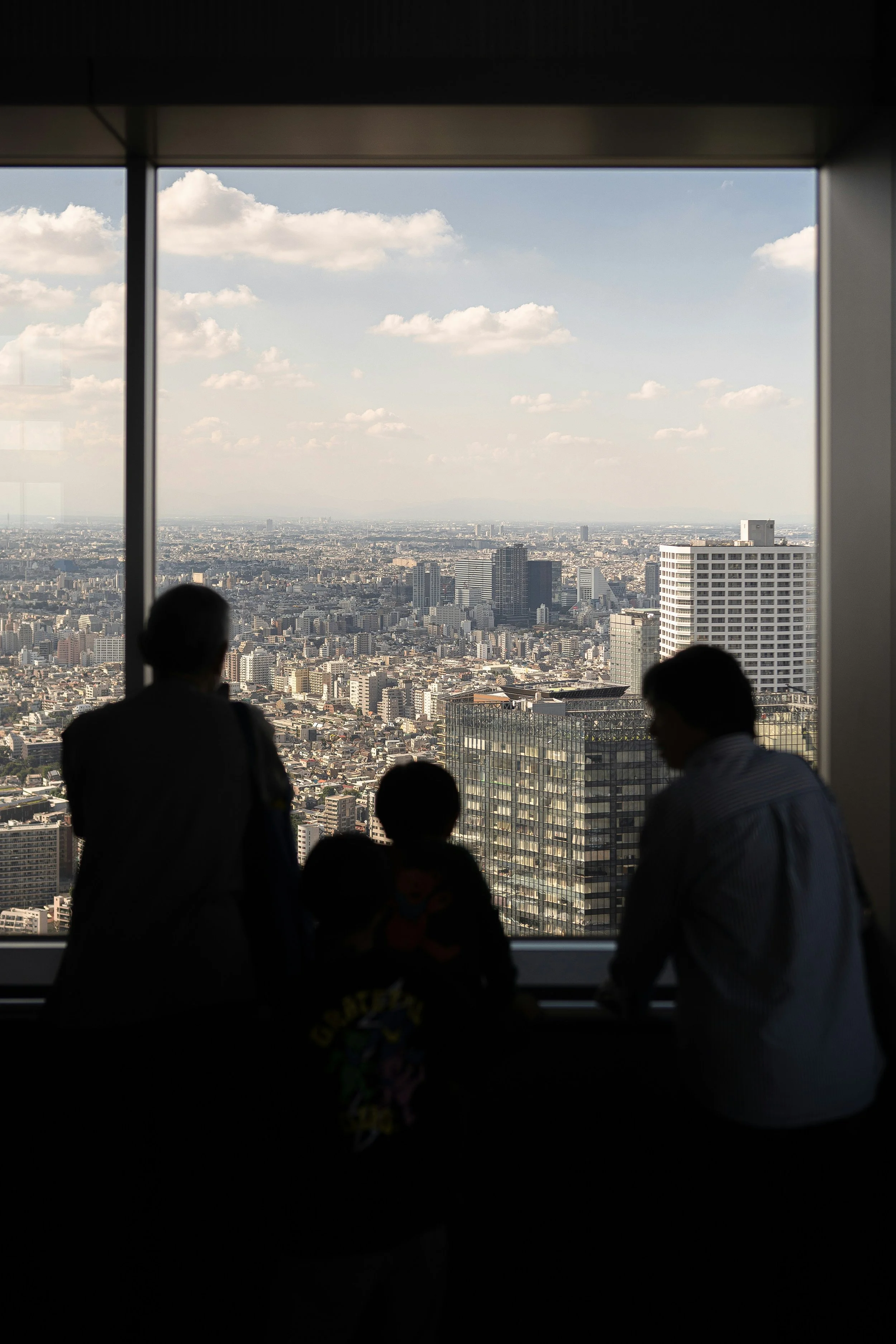 Silhouetted figures of three people looking out of a large window at a city skyline of Tokyo with tall buildings and a partly cloudy sky.