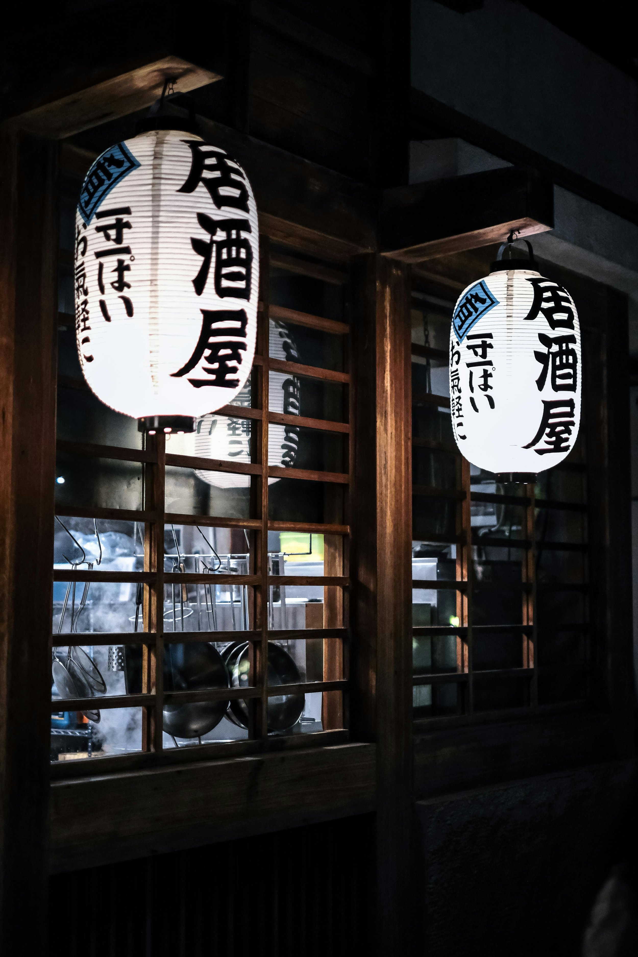 Japanese paper lanterns hanging in front of a wooden window, with kitchen utensils visible inside.