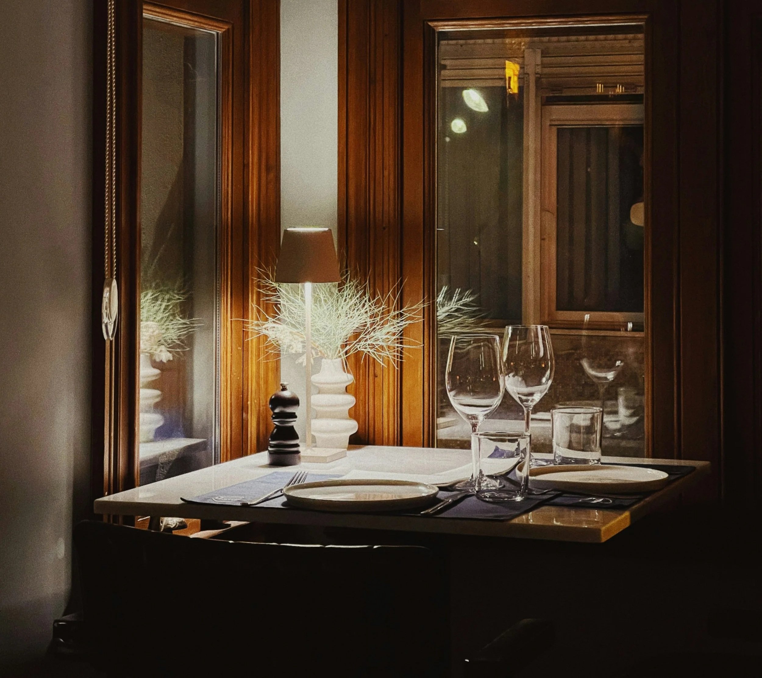A cozy dining table set near a window with wooden frames, decorated with a table lamp, a vase with decorative branches, and surrounded by wine glasses and plates in a warm, softly lit room.