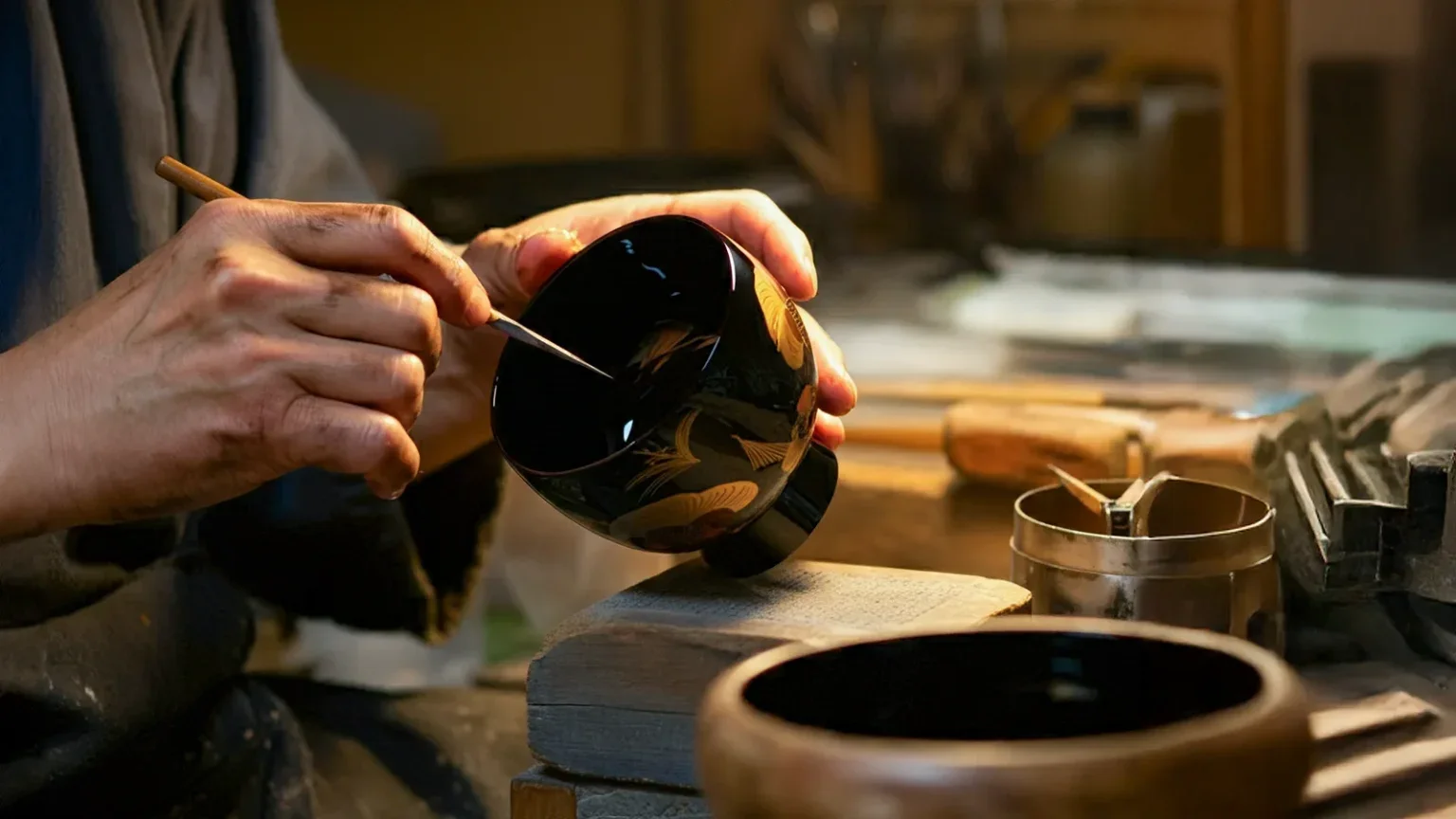 A person painting a black urushi bowl with gold design in a workshop. Japanese artisan.