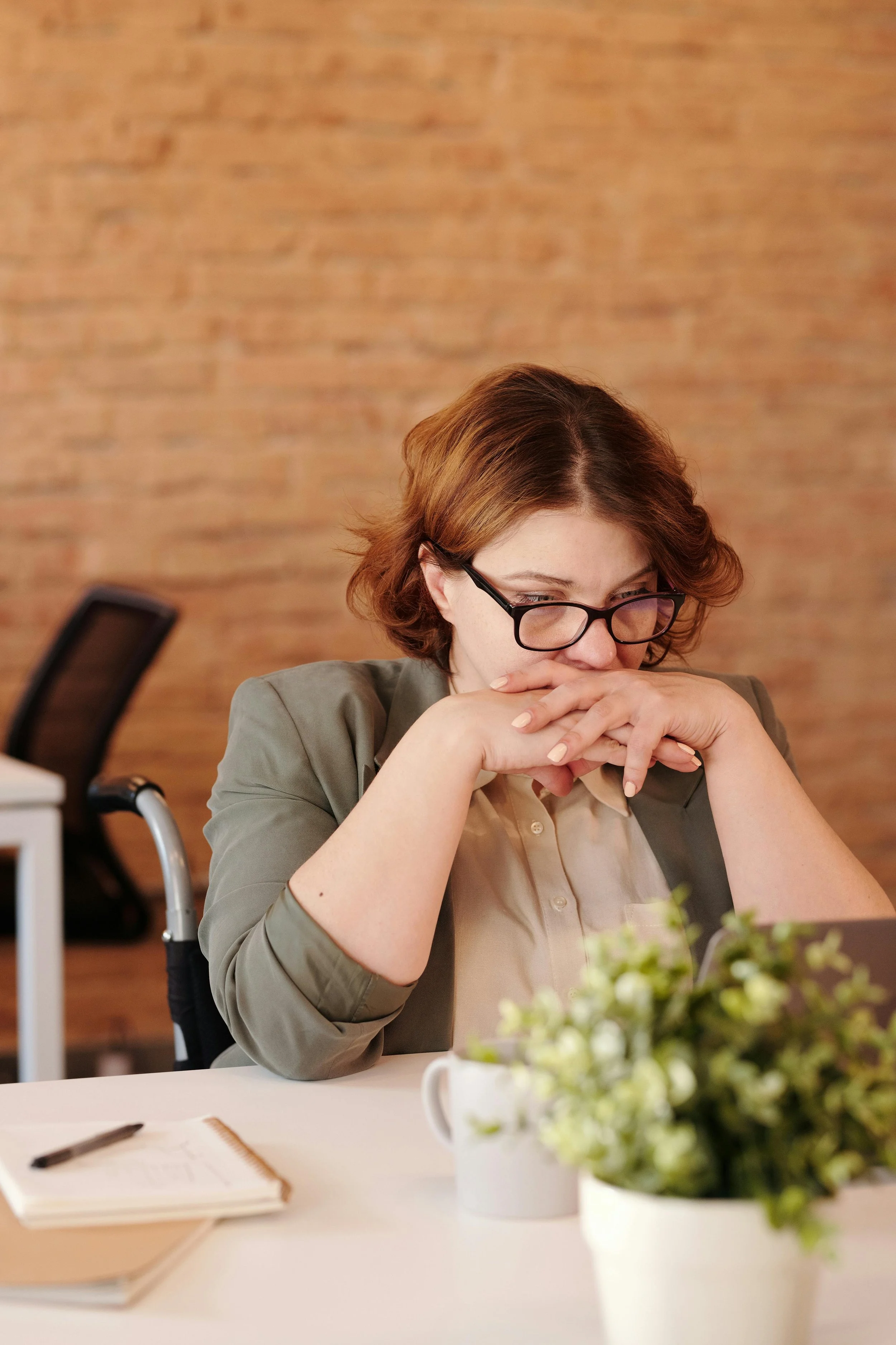 A woman with short red hair and glasses, wearing a gray blazer and beige shirt, sitting at a desk with a notebook, a pen, a mug, and a potted plant, appearing contemplative in an office with a brick wall background.