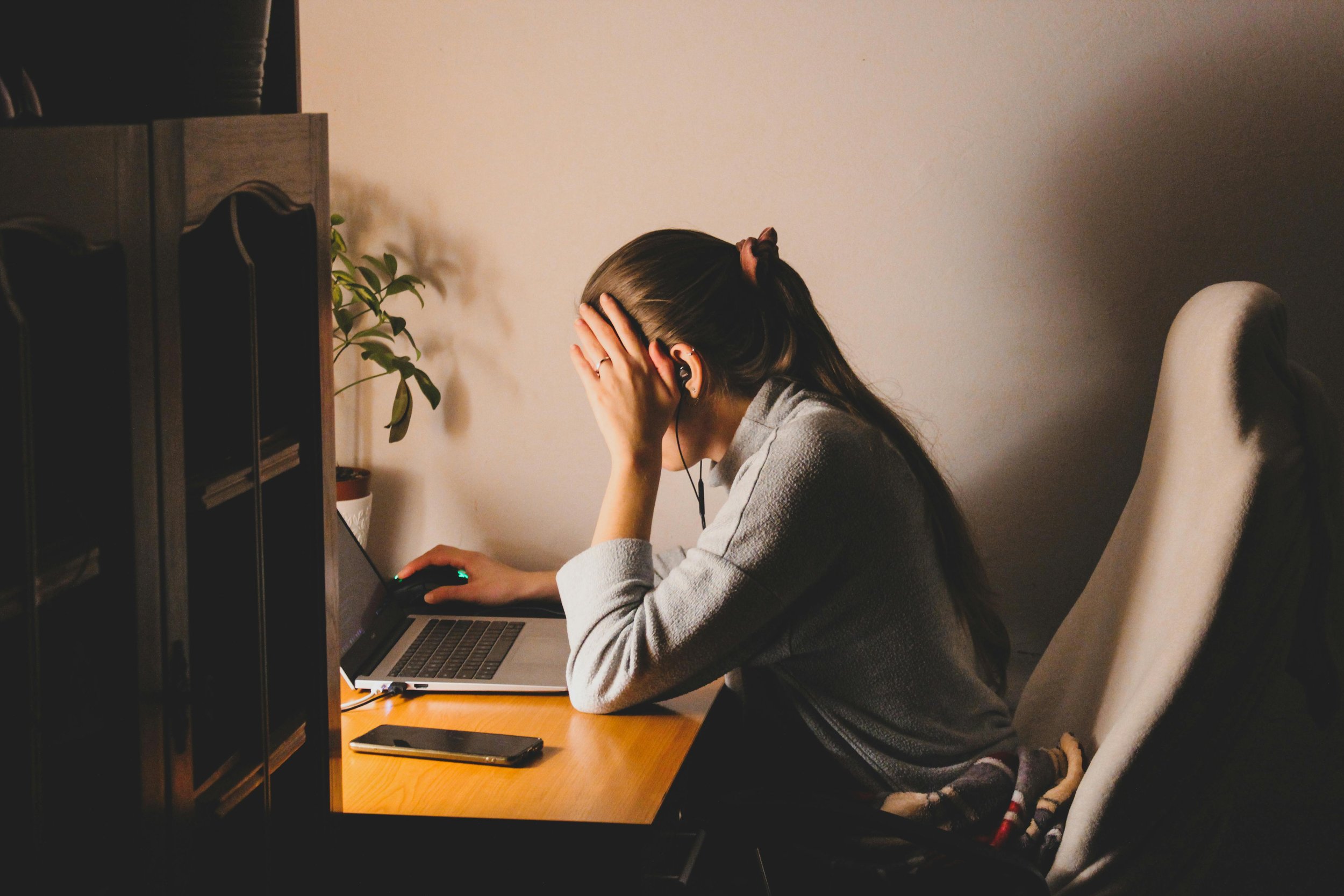A woman sitting at a desk with her head in her hand, using a laptop, wearing a gray sweater and earphones, in a dimly lit room.