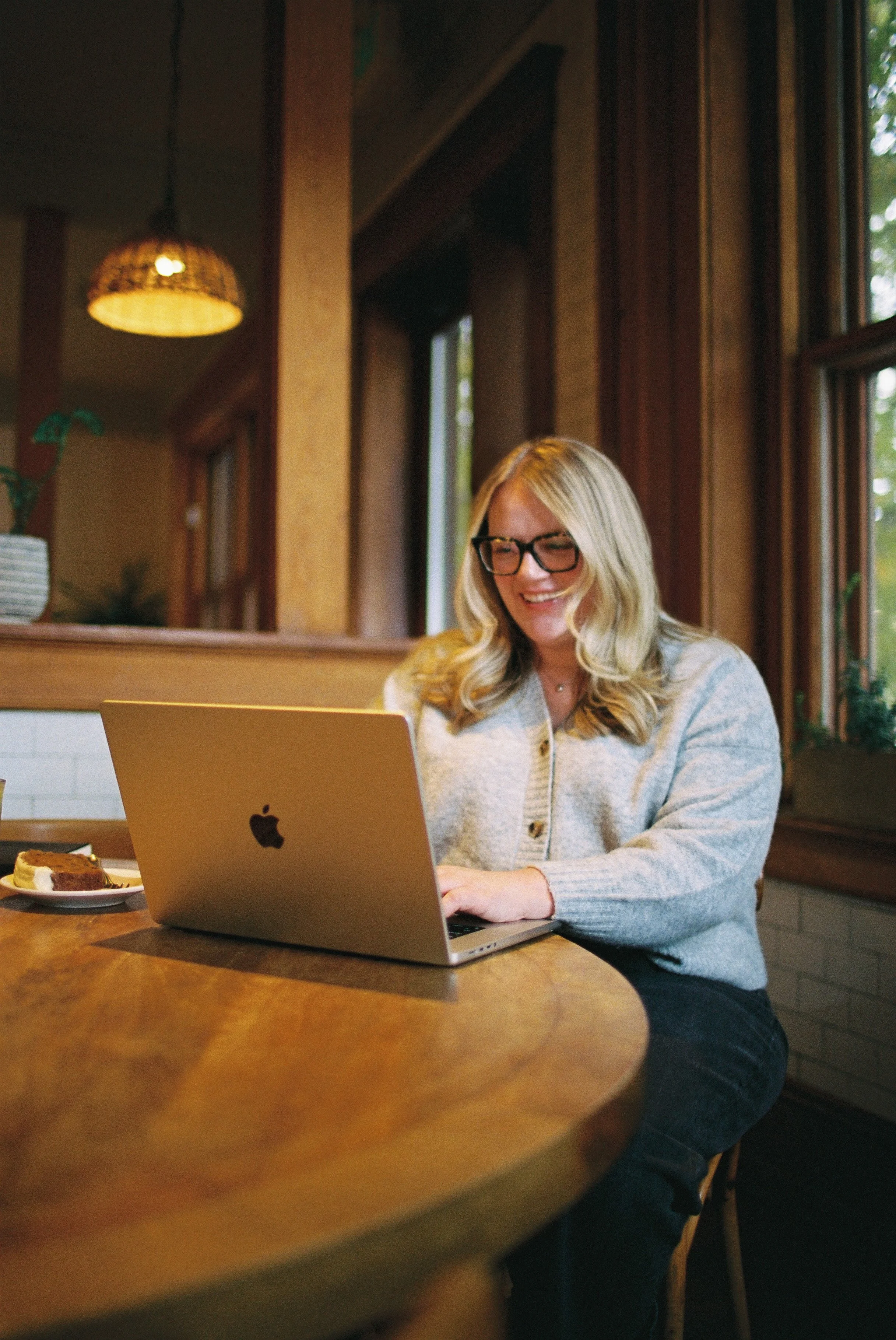 A woman with blonde hair and glasses working on a laptop at a wooden table in a cozy, wood-paneled room with windows and a potted plant in the background.