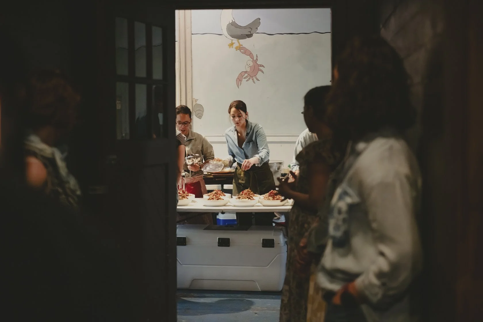 People preparing food behind a counter in a kitchen or restaurant, with a colorful cartoon fish and crab painted on the wall in the background.