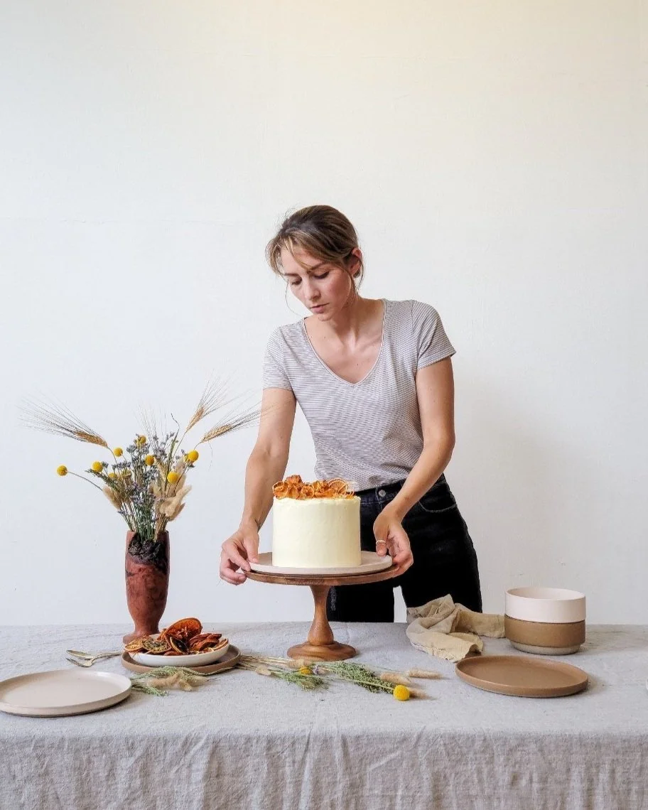 A woman arranging a white frosted cake decorated with caramel and flowers on a round cake stand on a table. The table is set with plates, a vase with dried flowers, and other tableware.