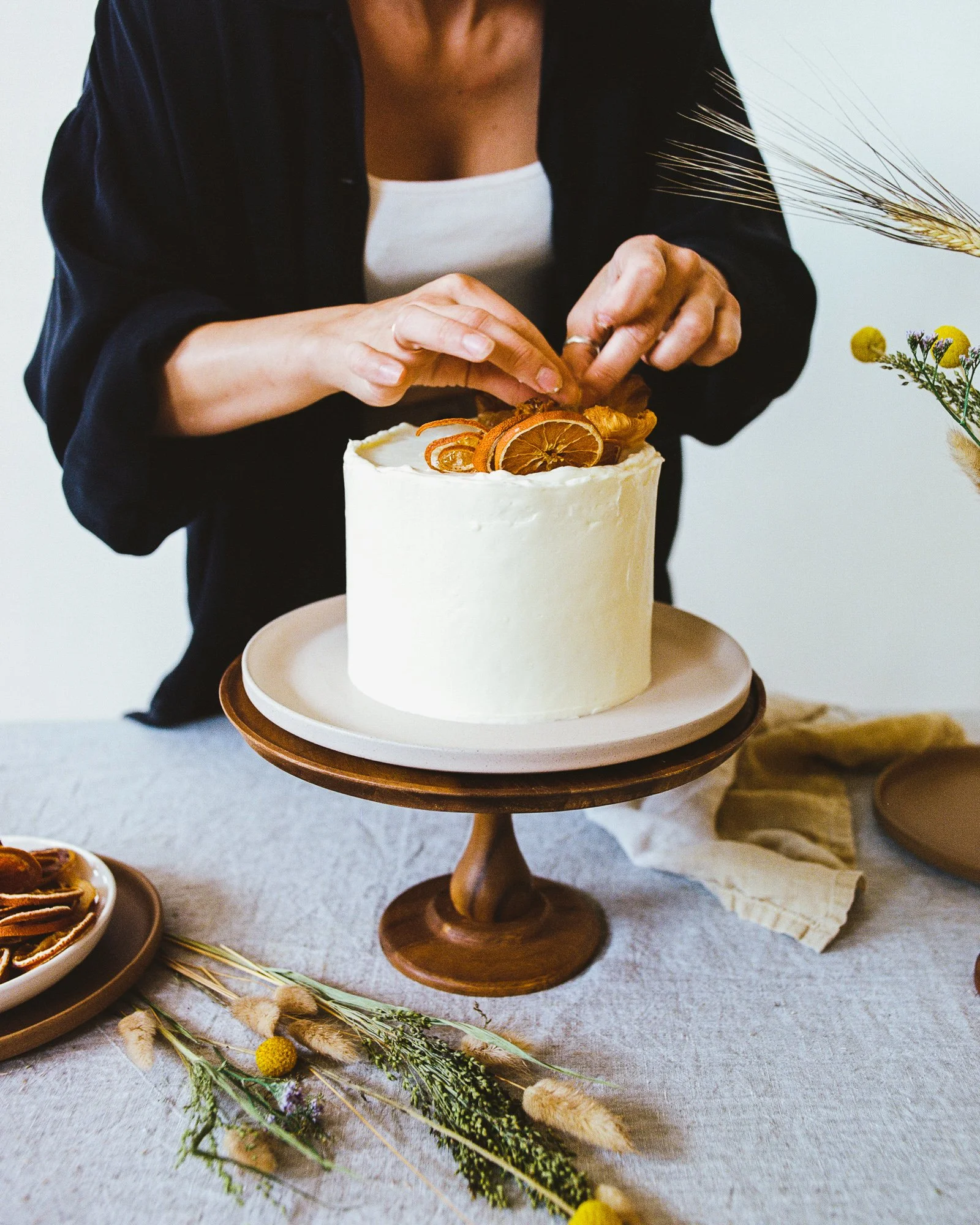 Person decorating a white frosted cake with dried orange slices, on a cake stand, at a decorated table.