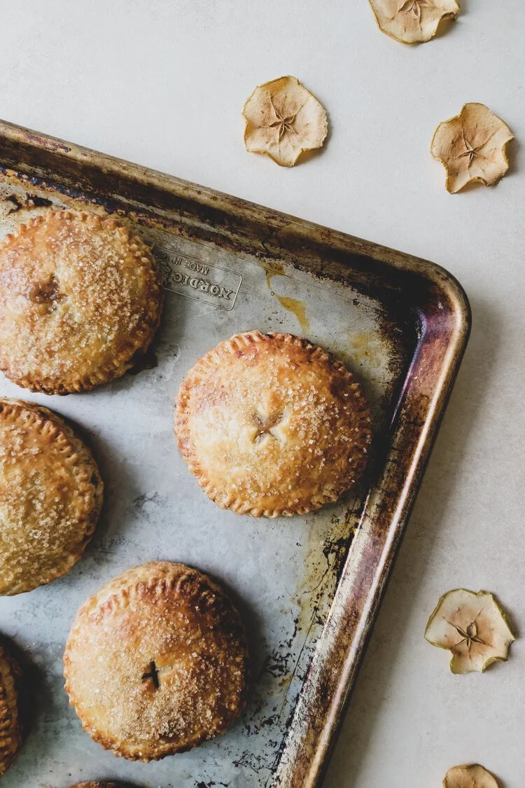 Baked apple hand pies sprinkled with sugar, on a baking sheet, with dried apple slices on a white surface.