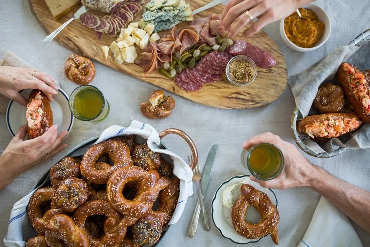 A charcuterie board with assorted cheeses, cured meats, and pickles, surrounded by plates of pretzels, dipping mustard, and beer, with hands reaching for food.