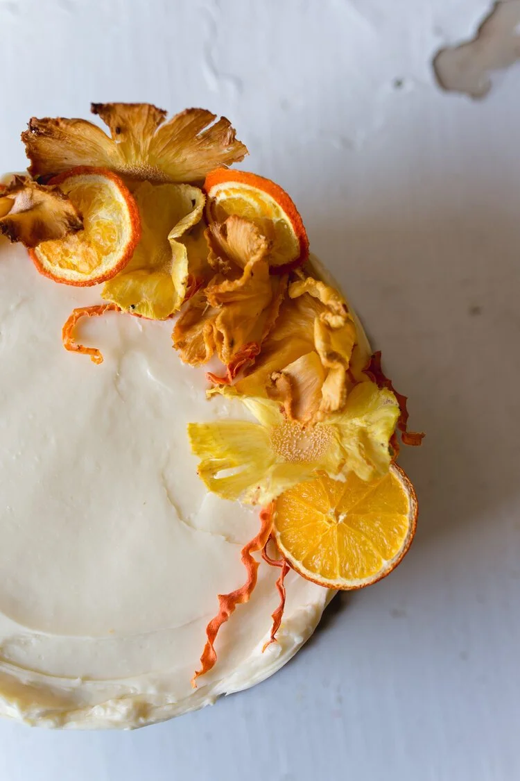 Decorative cake topped with dried pineapple, orange slices, and candied citrus peel on a white surface.