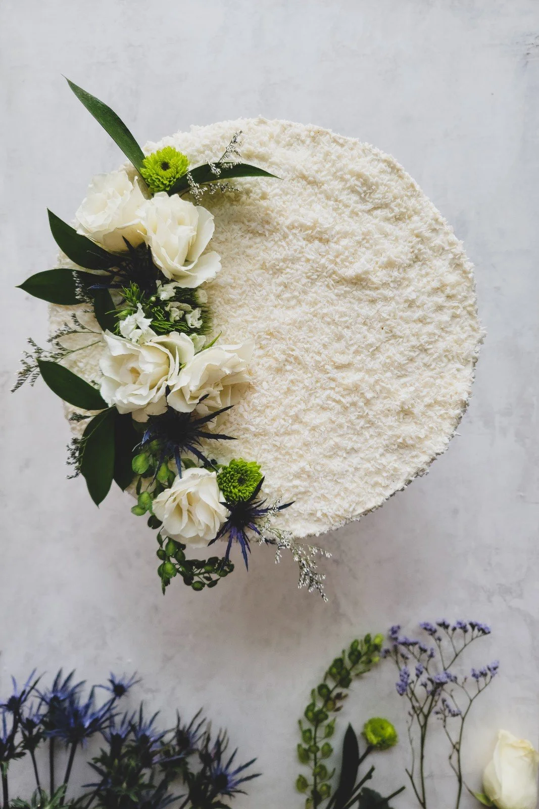 Round white textured cake decorated with white roses, green leaves, and small purple and green flowers.