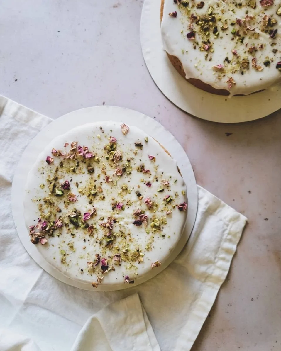 Two round cakes with white icing and sprinkled with crushed pistachios and dried rose petals, placed on white parchment paper on a light-colored surface.