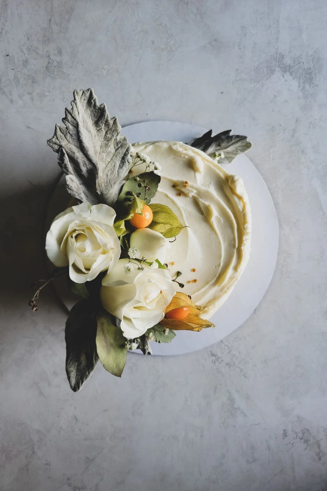 A cake decorated with white flowers, gray leaves, and small orange berries, viewed from above against a light gray background.