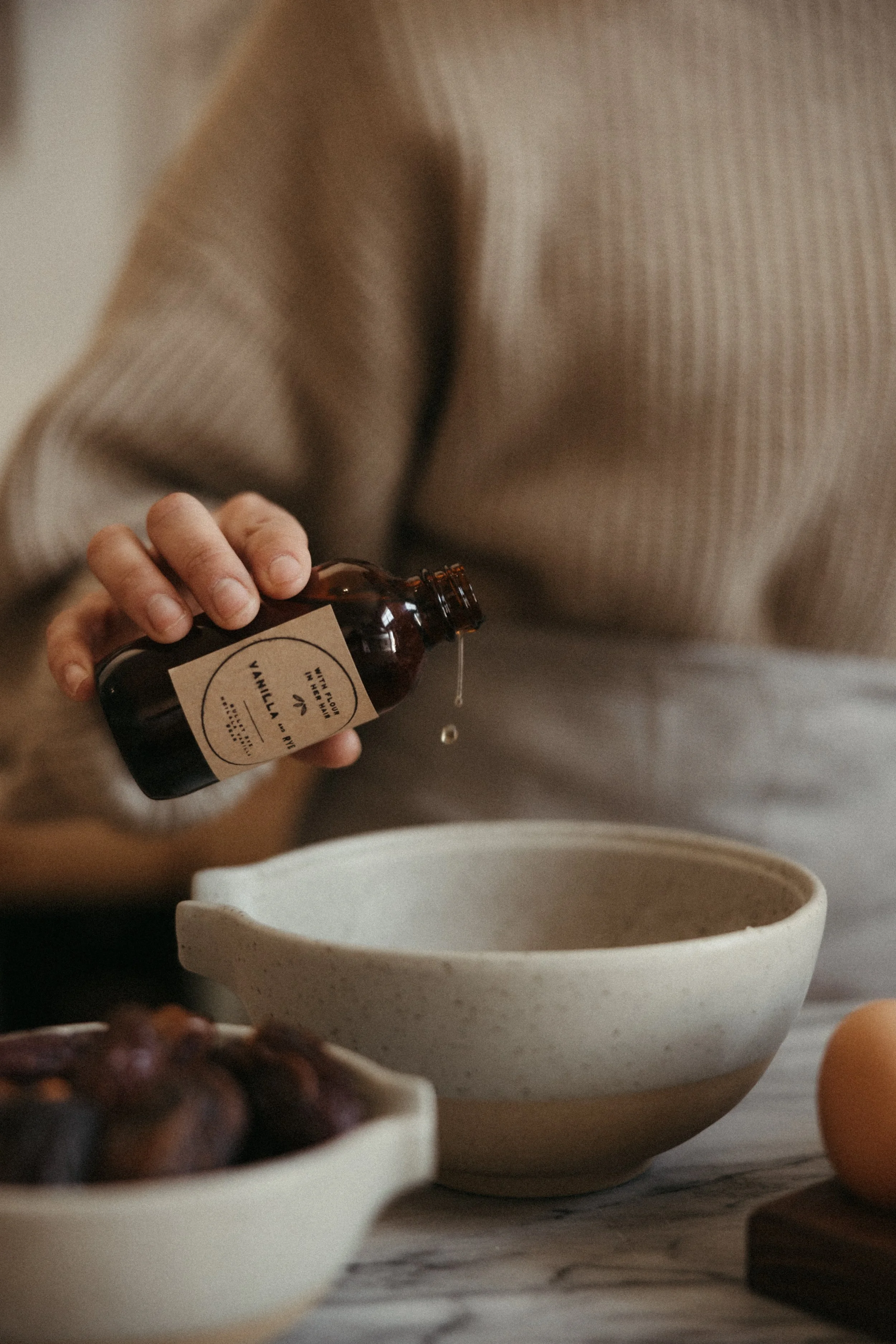 Person pouring liquid from a brown bottle labeled 'Vanilla' into a beige bowl on a kitchen counter.