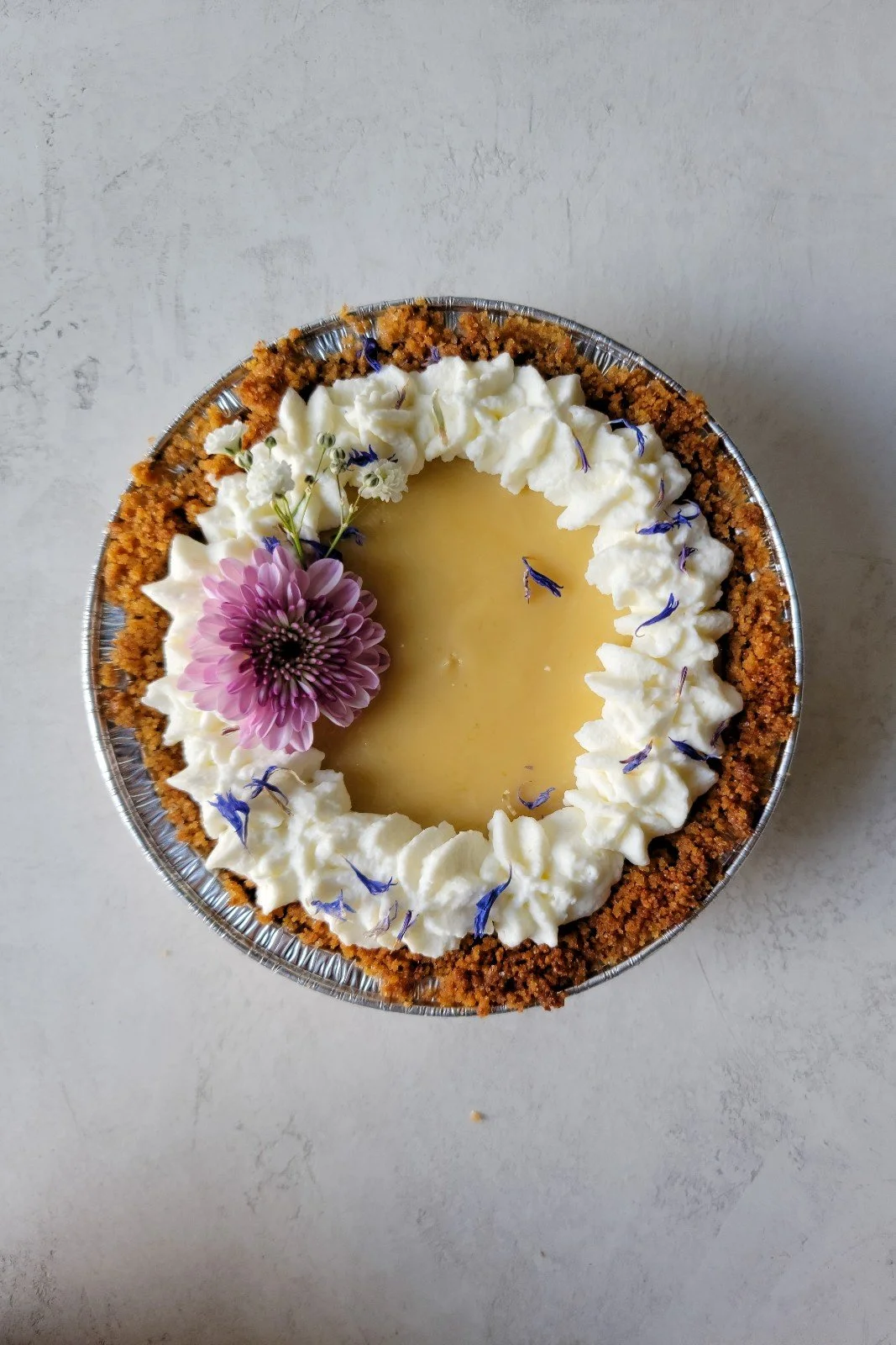 A dessert tart with whipped cream, edible flowers, and a crumbly crust in a round aluminum dish, on a light surface.