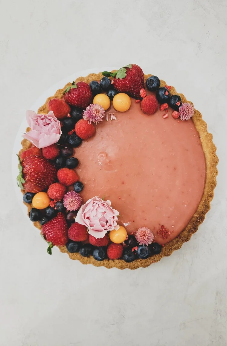 A round berry tart with a graham cracker crust, topped with strawberries, raspberries, blueberries, yellow berries, pink flowers, and a pink icing decoration.