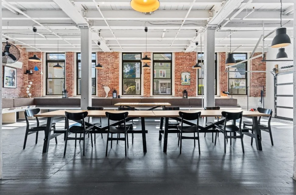 A modern, spacious meeting or dining area with a long wooden table surrounded by black chairs. The room features exposed brick walls, large windows allowing natural light, pendant light fixtures, and a garage-style door. Decor includes artwork on the walls and a bench against the brick wall.