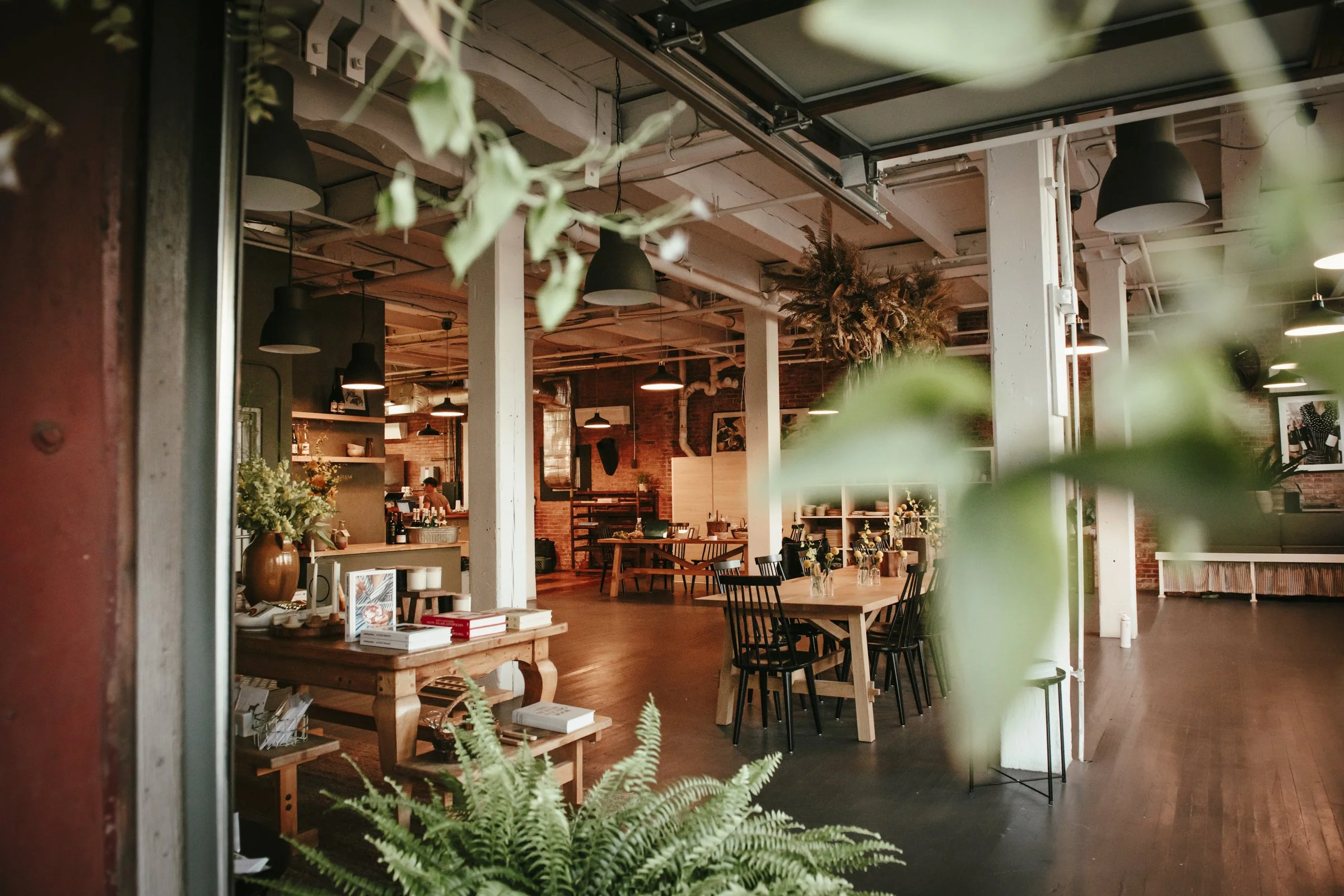 Indoor cafe or restaurant with wooden tables and black chairs, brick walls, green plants, and hanging light fixtures.