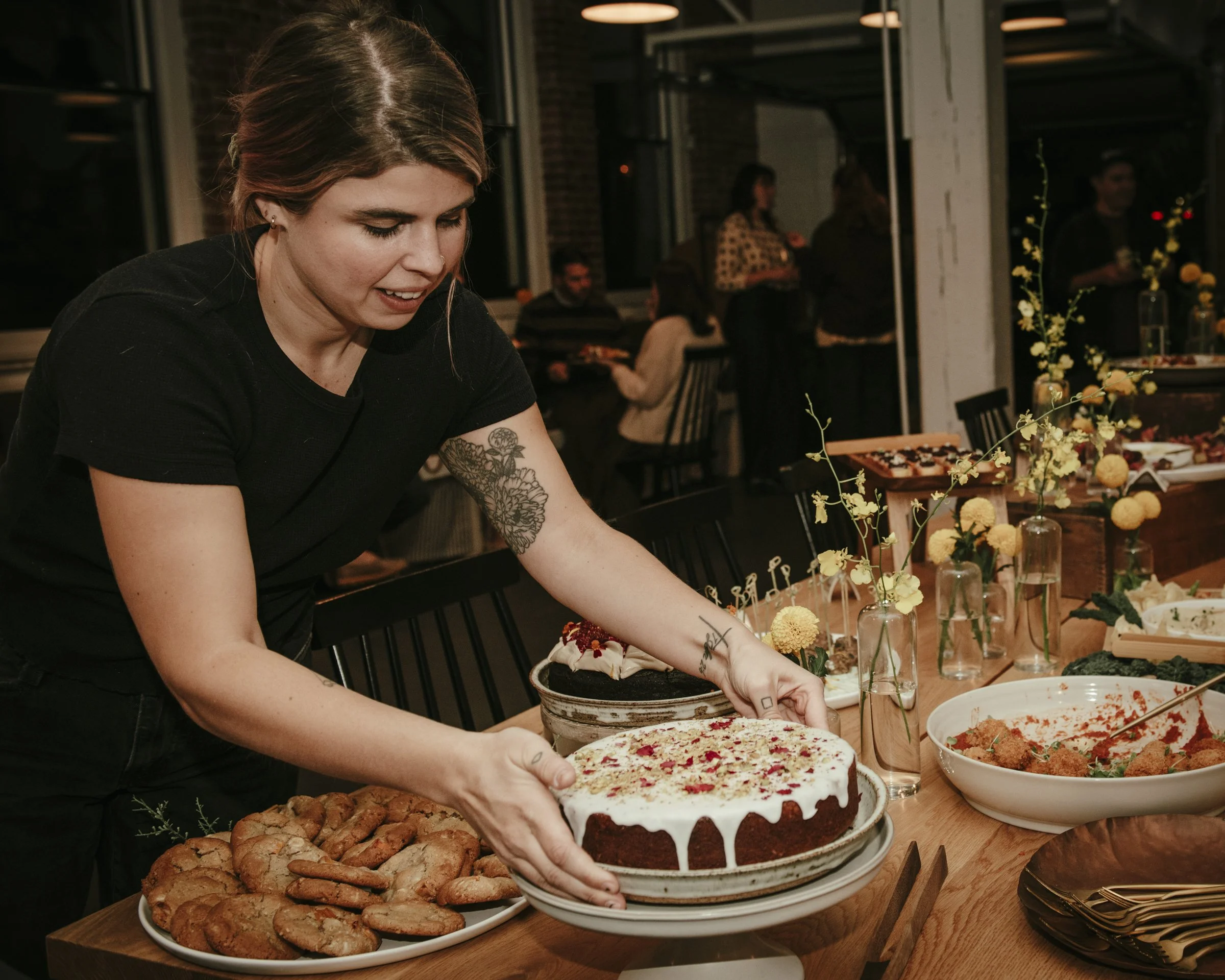 A woman with tattoos on her arm is placing a decorated birthday cake on a table at a gathering.