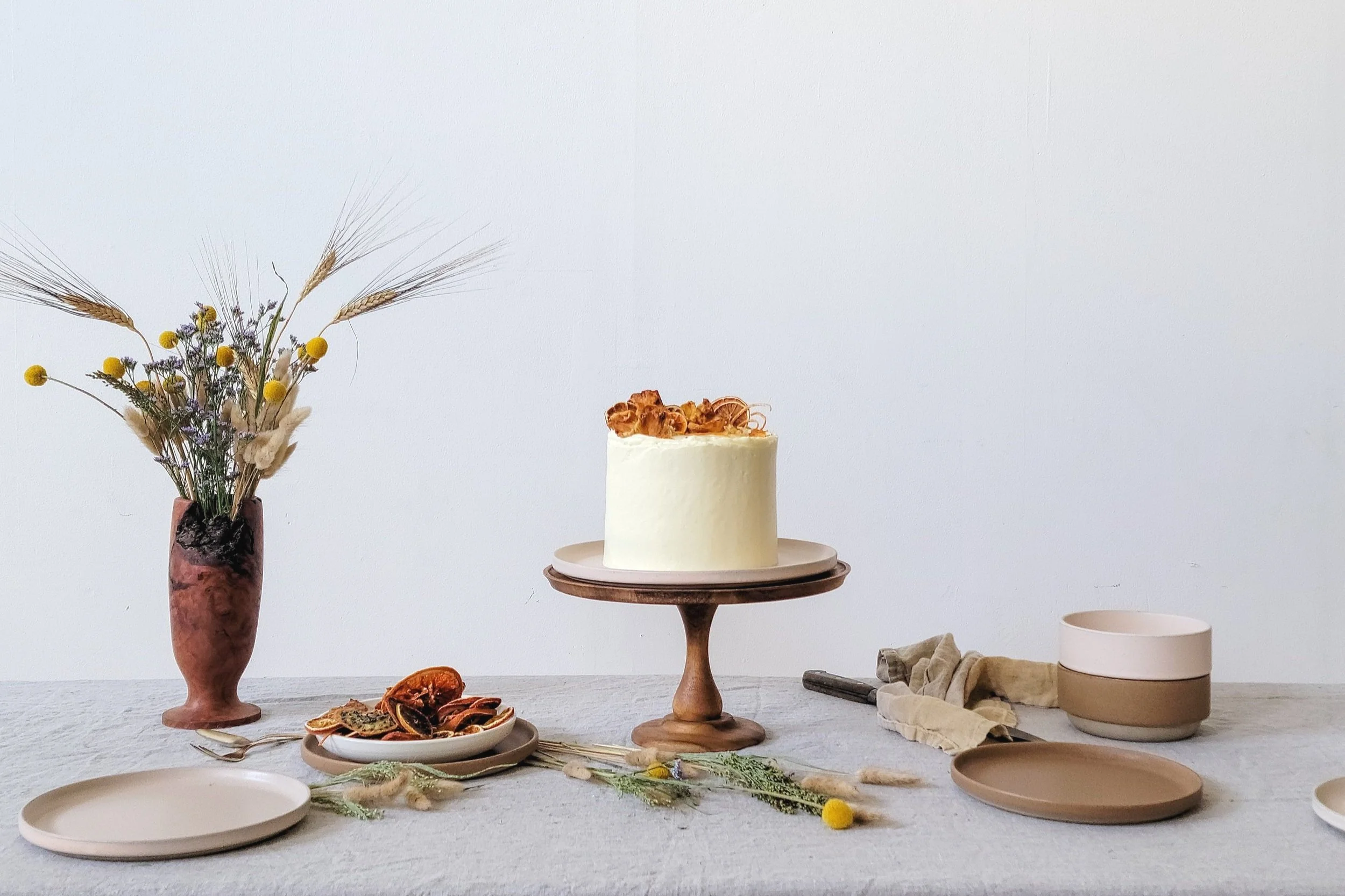 A white frosted cake on a wooden cake stand, decorated with caramelized walnuts or similar topping, is centered on a table. To the left, a tall vase with dried flowers and wheat stalks. In front, a plate with dried fruit or bark-like slices. To the right, a beige and pink bowl, a spatula, and some folded fabric are visible, with a neutral tablecloth covering the table.