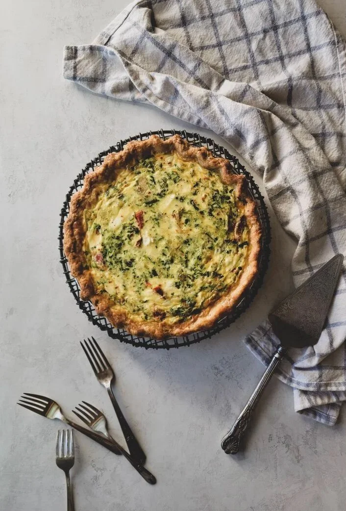 A vegetable quiche on a black wire cooling rack, with a scalloped pastry crust, set on a light-colored surface. A gray and white checked cloth and a metal pie server are nearby, along with four forks on the surface.
