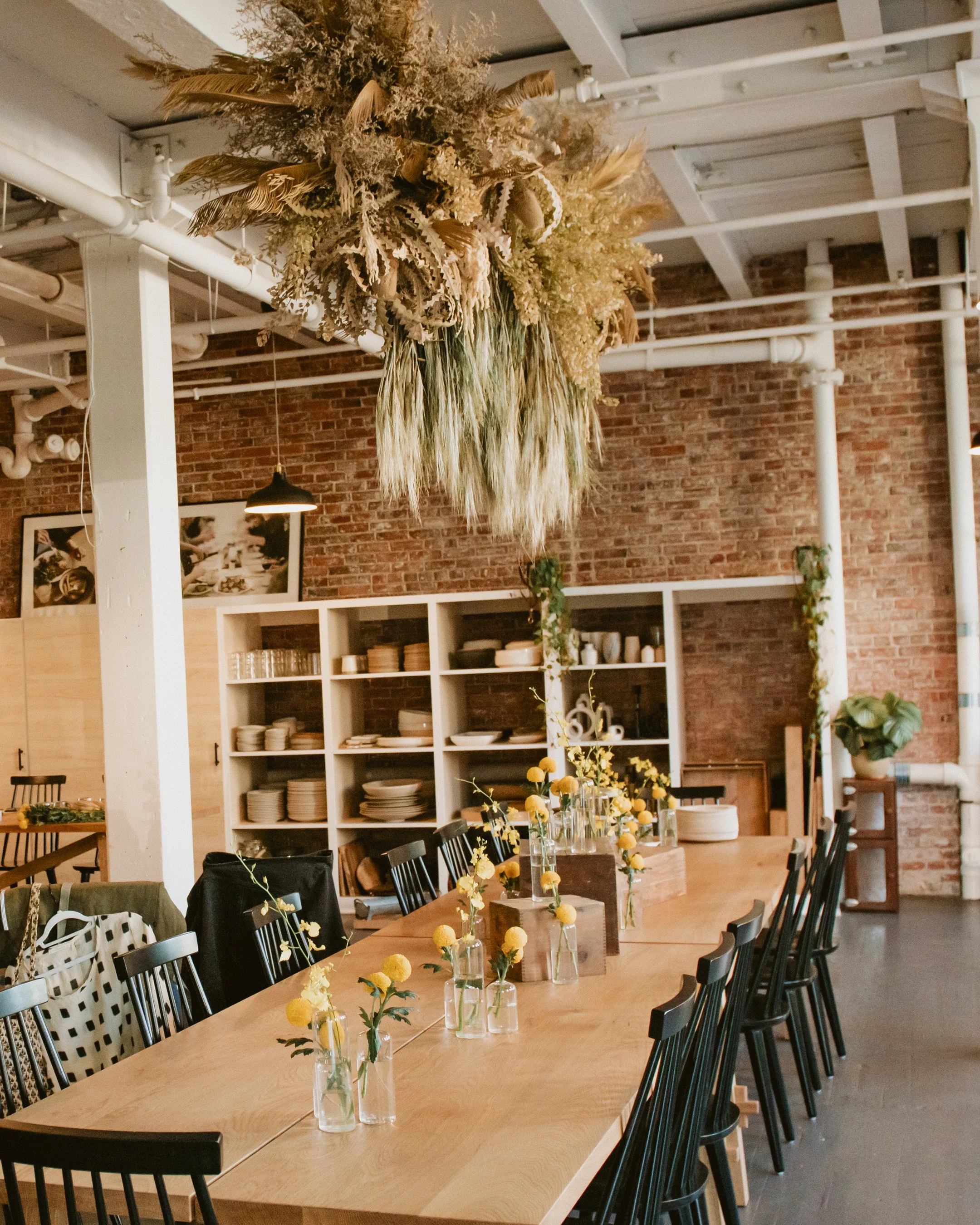 A dining room with a long wooden table decorated with small vases of yellow flowers. Overhead, a large dried floral arrangement hangs from the ceiling. The room has a brick wall, white shelves filled with dishes, and black chairs around the table.