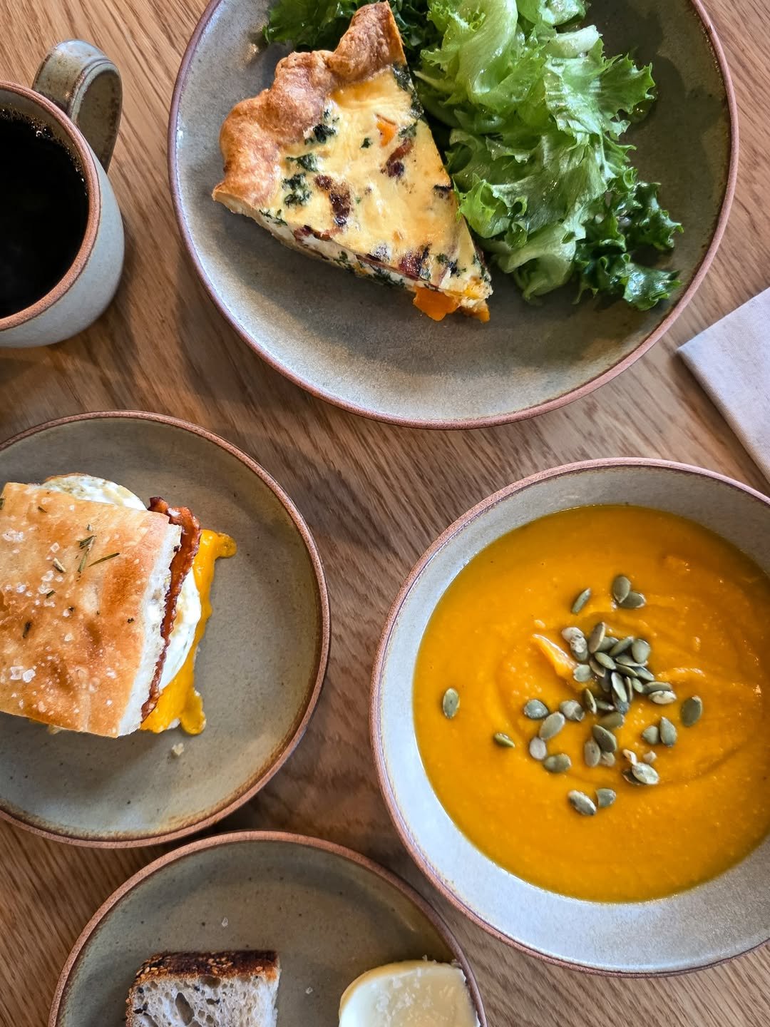 A table with bowls of pumpkin soup, salad, quiche, and a breakfast sandwich with a drink.