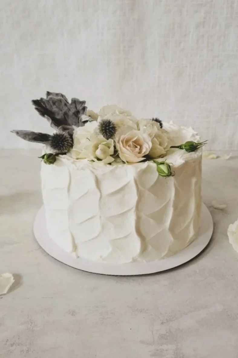 White wedding cake decorated with white and pale pink flowers and gray foliage on top, placed on a white cake board.