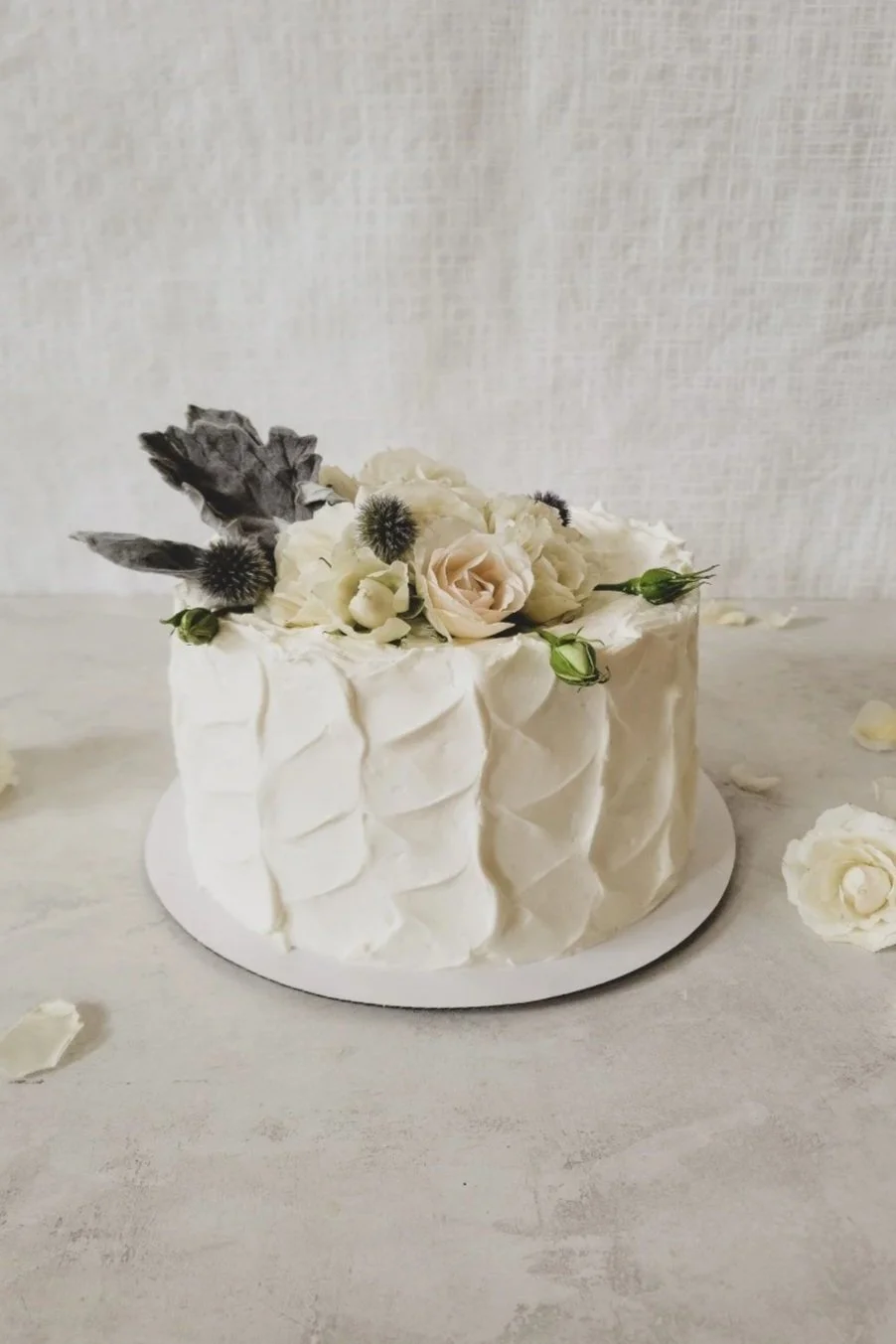 White cake decorated with white flowers and gray foliage on top, with a textured pattern on the sides, on a white cake board.