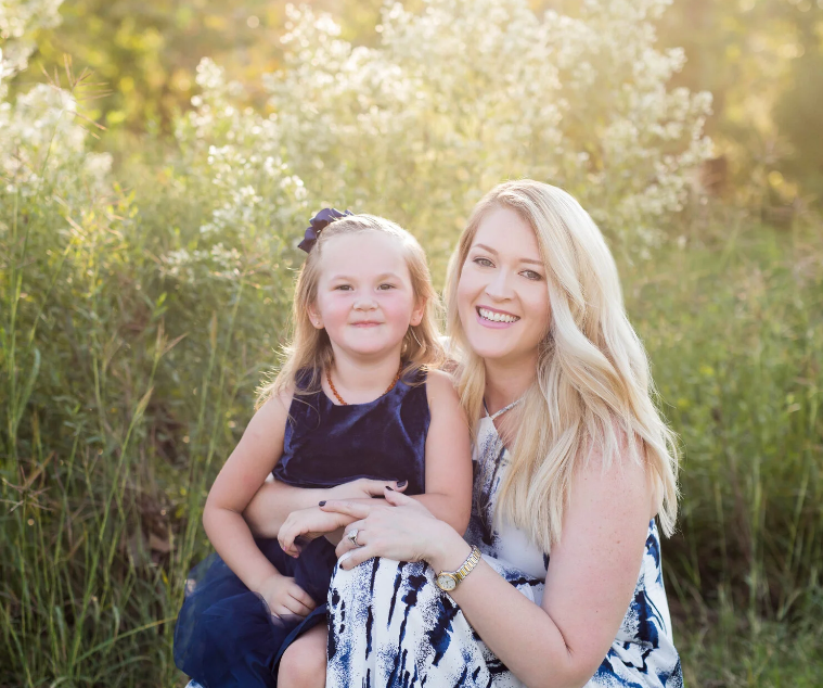 A woman and a young girl sitting outdoors in a natural setting with tall grass and flowering plants, both smiling at the camera.