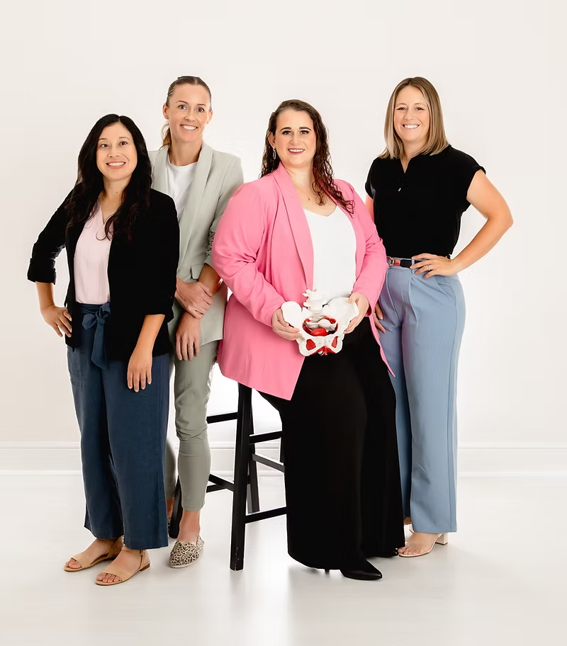 Four women standing together against a white background, with one holding a pelvic model.