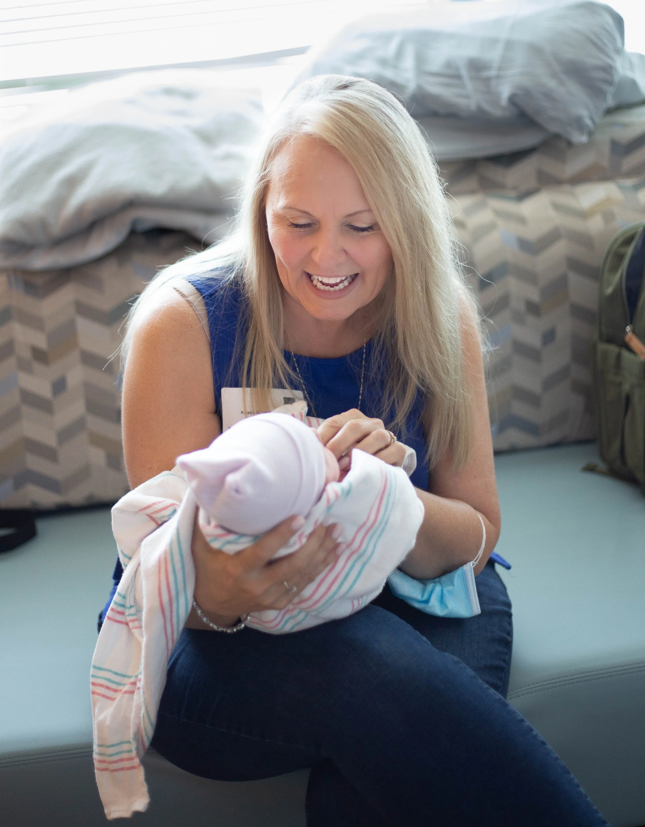 A woman with long blonde hair holding a newborn baby wrapped in a hospital blanket, smiling and looking at the baby in a hospital room.