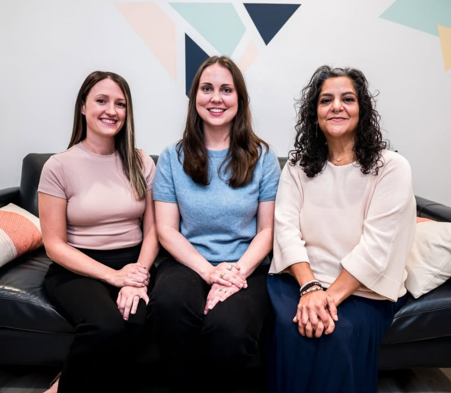 Three women sitting on a black couch in a room with geometric wall art, smiling at the camera.