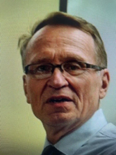 A middle-aged man wearing glasses and a dress shirt, looking surprised or confused.