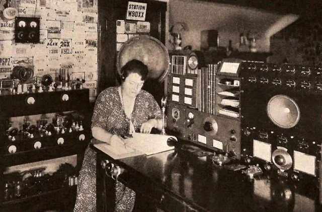 Black and white photo of a woman standing at a desk with vintage radio and recording equipment, in a radio station or broadcast studio
