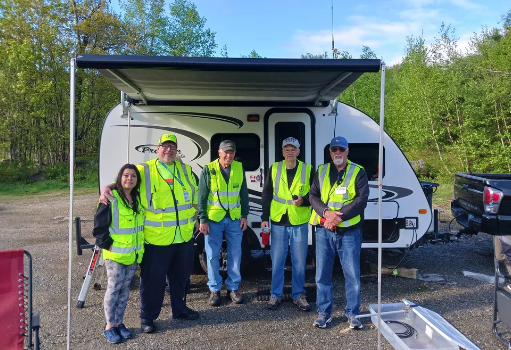 Five people standing in front of a white trailer with a pop-up awning, wearing neon yellow safety vests, in an outdoor setting with trees and a gravel ground.
